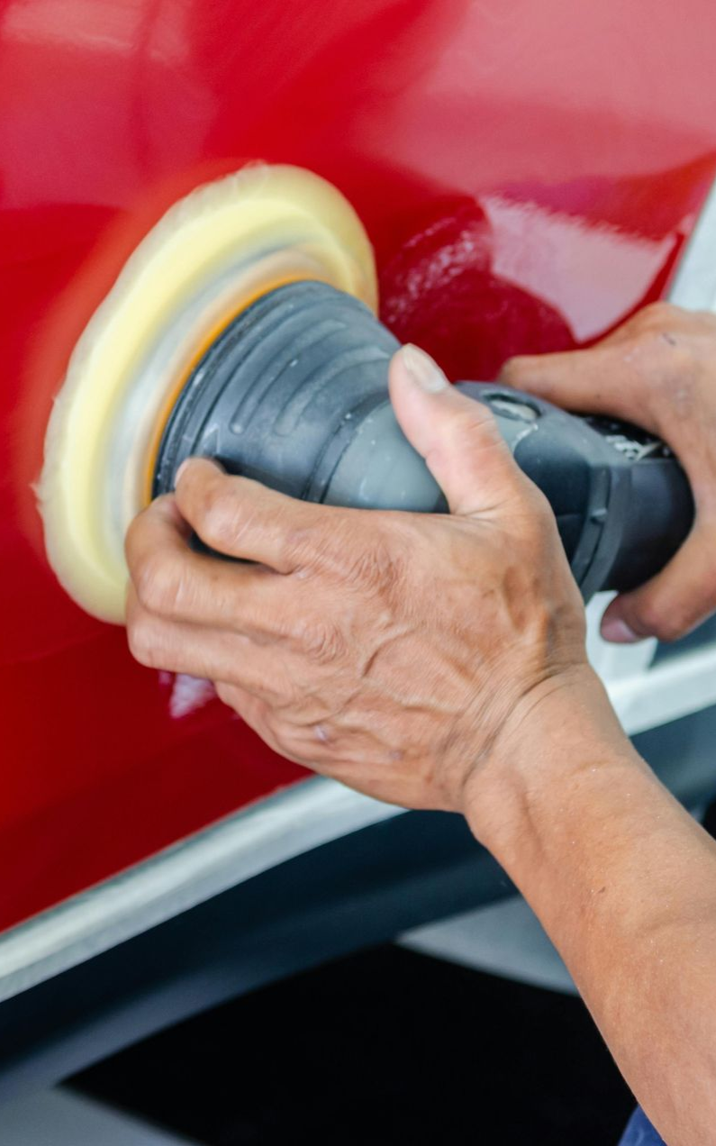 Person using a power buffer to polish a red car panel with a white polishing pad.