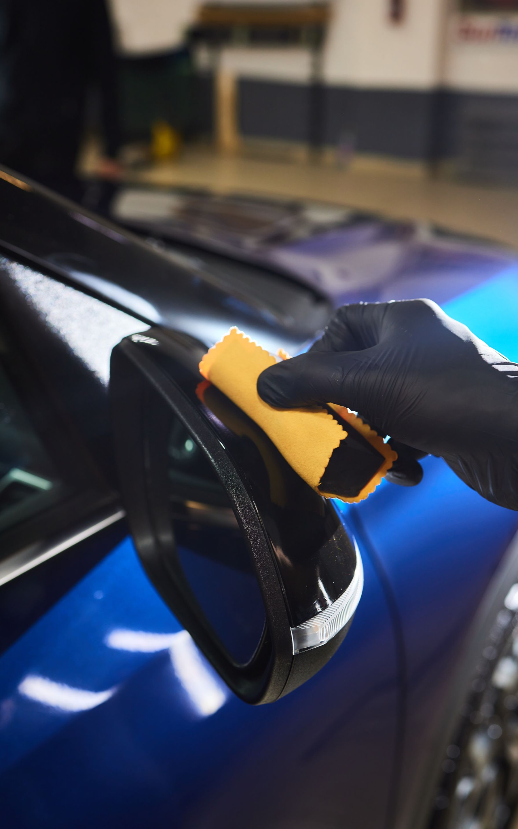 Gloved hand applies ceramic coating to a car's black side mirror in a workshop. Blue car visible.