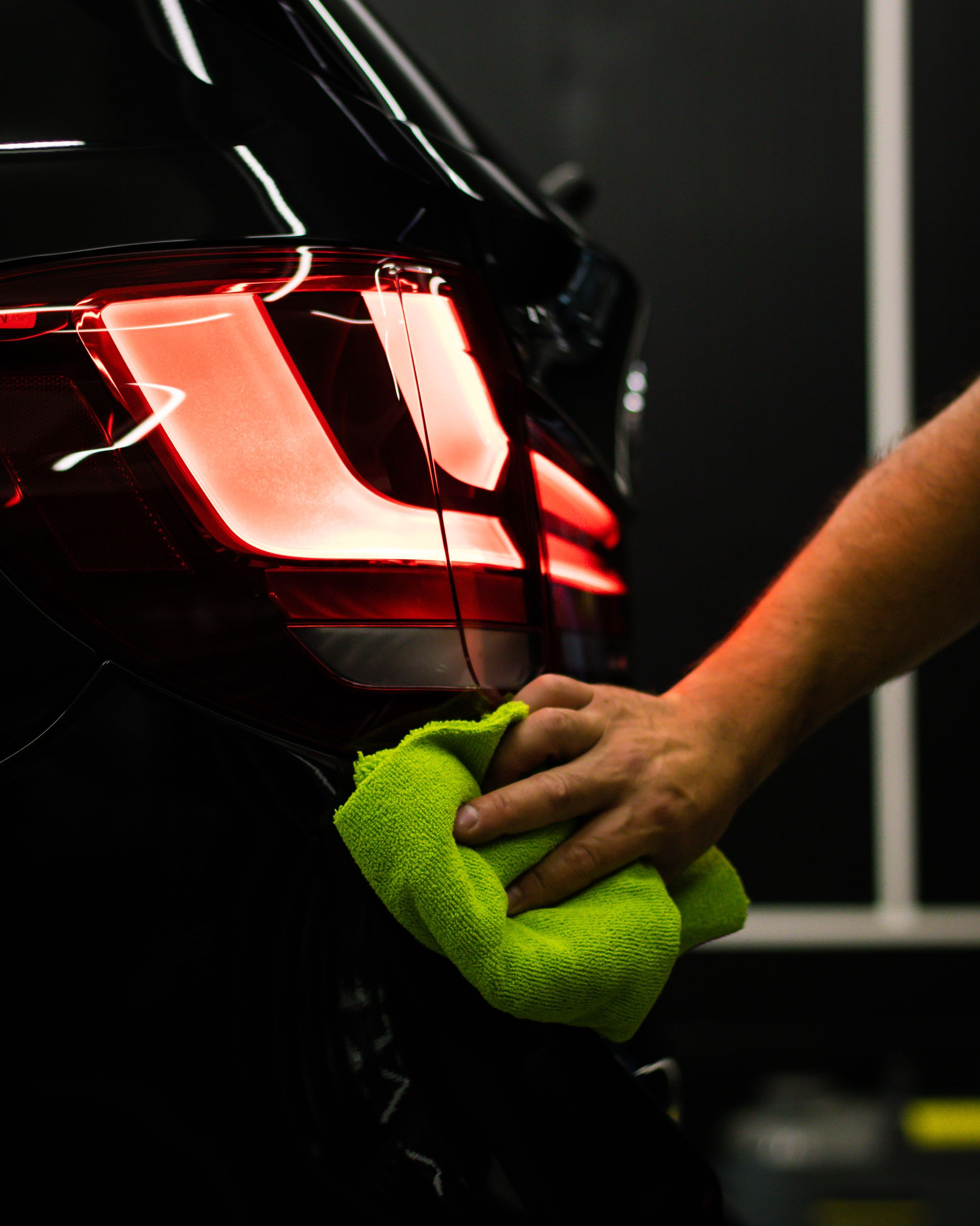 Person wiping a black car's taillight with a green microfiber cloth; dark background.