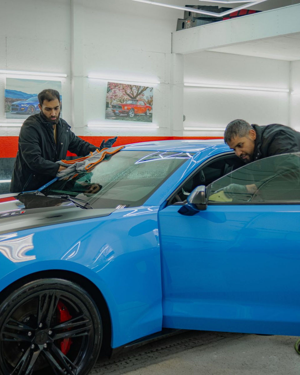Two men working on a blue car in a garage. One applies tint, other inspects inside.