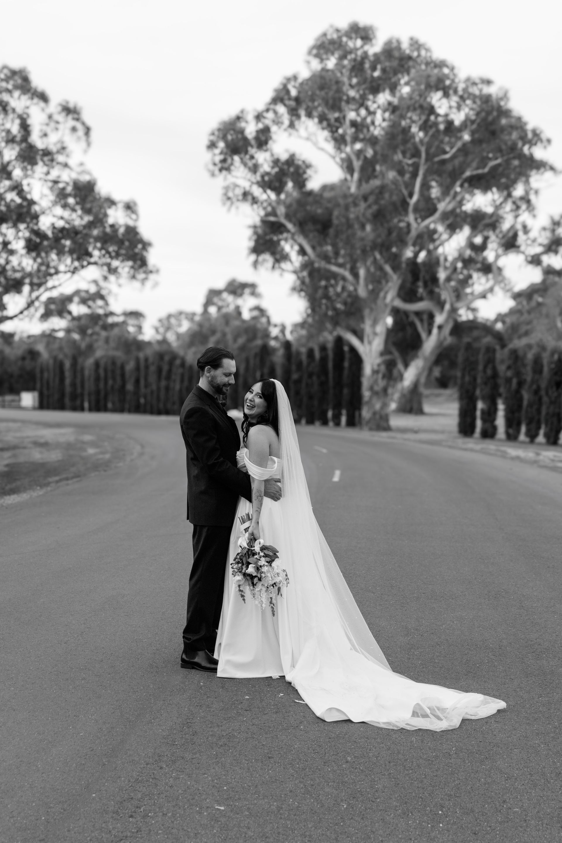 A black and white photo of a bride and groom standing next to each other on a road.