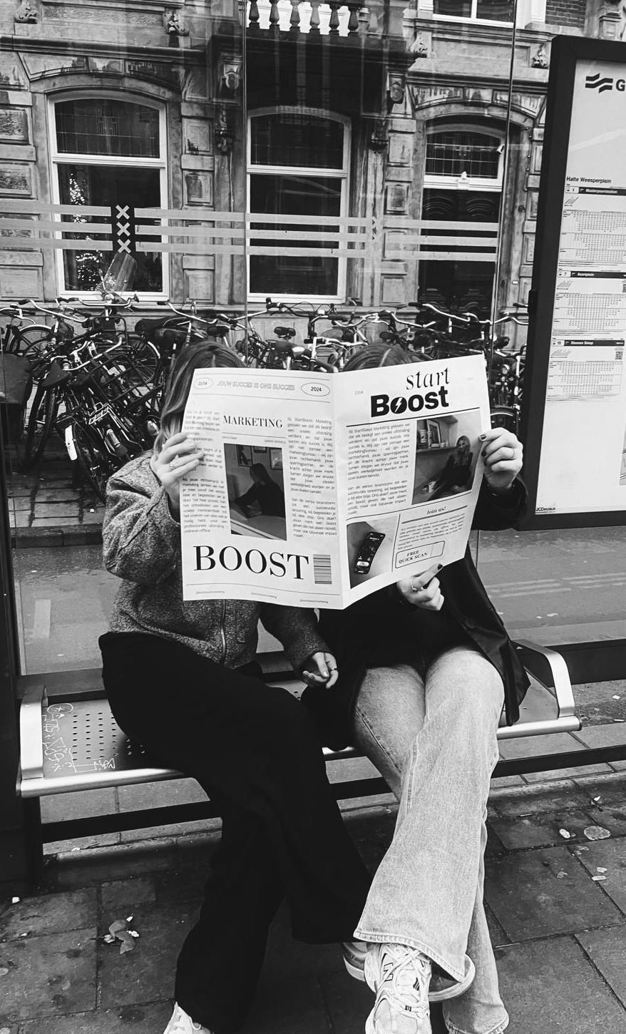 Twee mensen zitten bij een bushalte en lezen een krant.