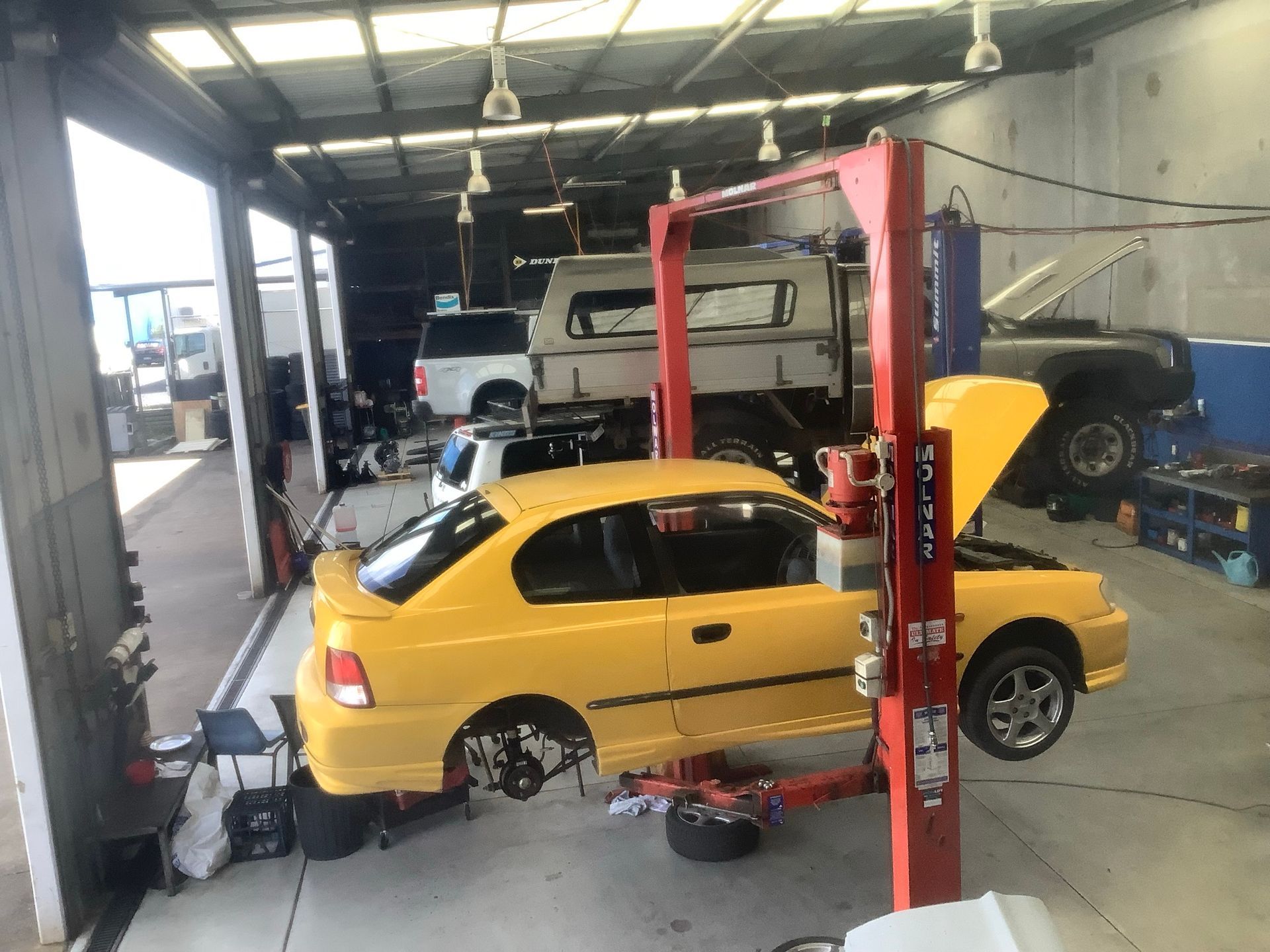 Yellow car on a lift in a mechanic's shop with hood open, other vehicles in the background.