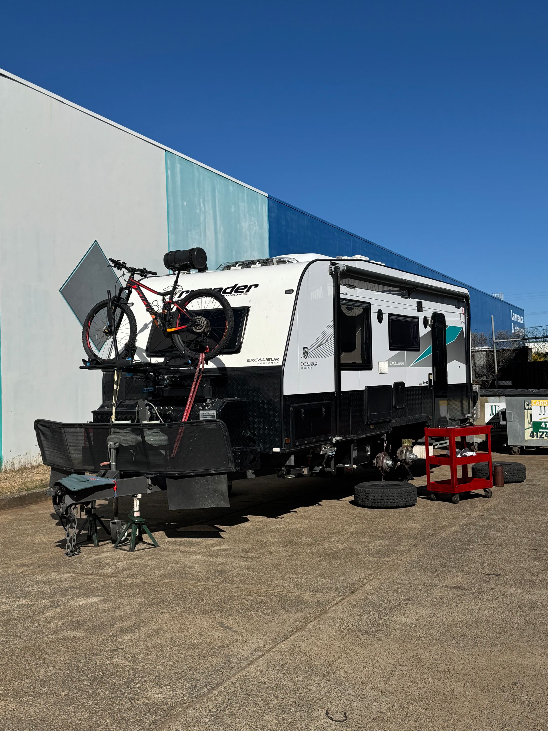 RV with bike on top, tracks instead of wheels, parked in front of a building on a sunny day.