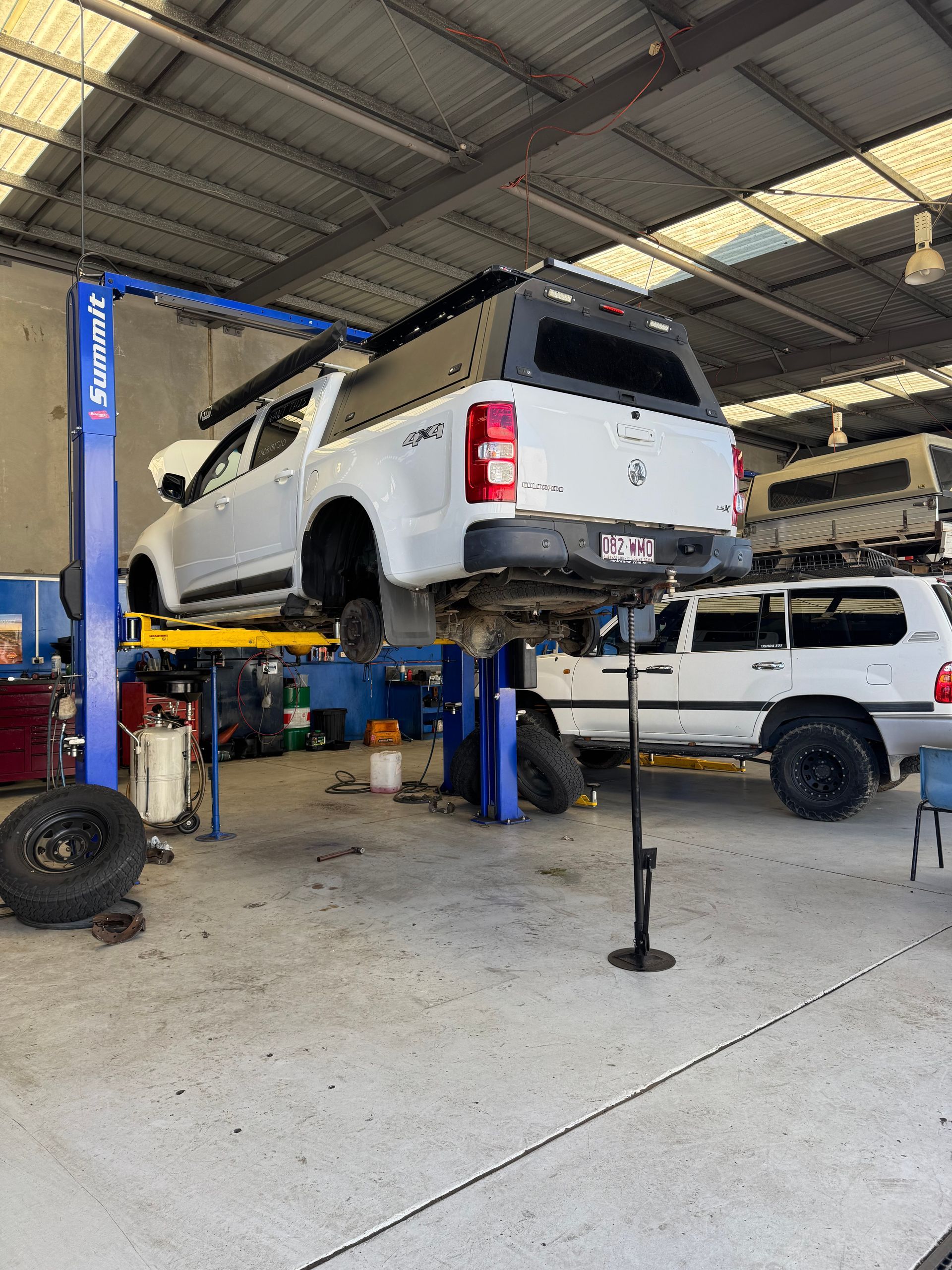 White truck on a lift in a garage, being worked on, with another white truck and equipment in the background.