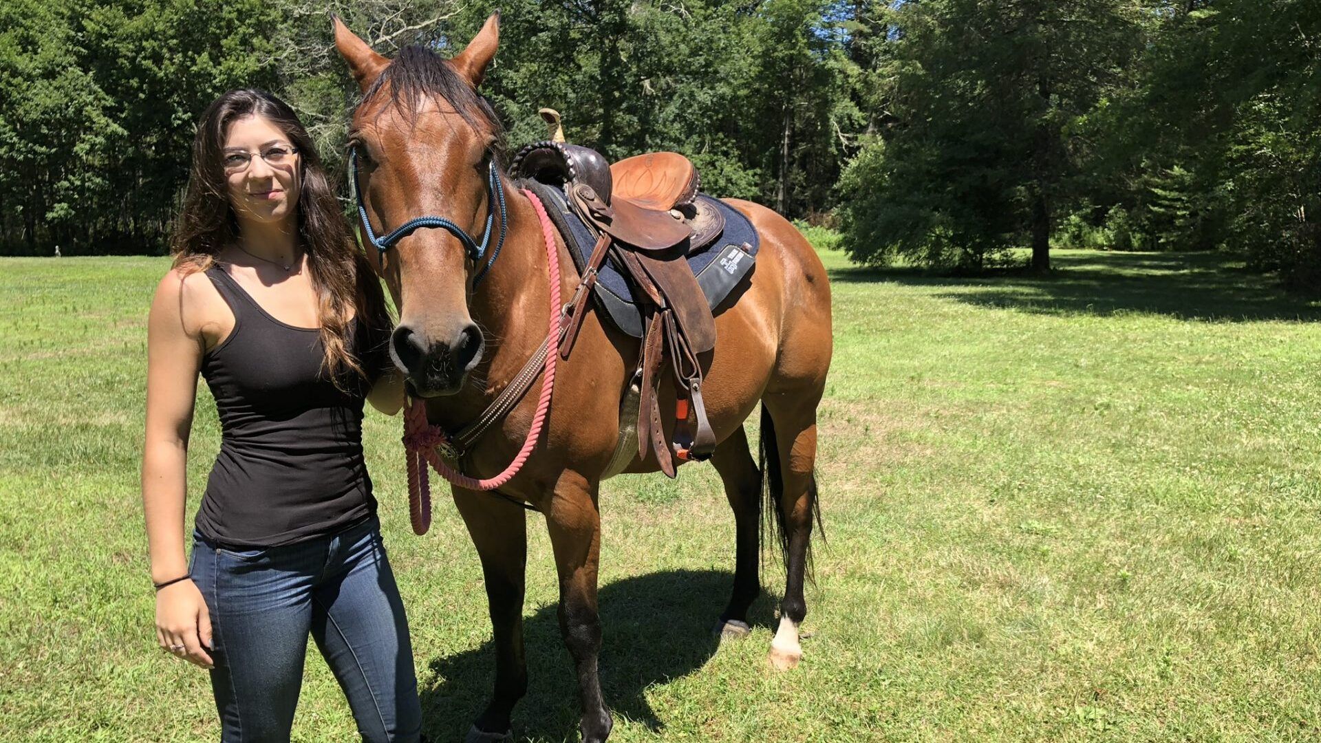 A woman is standing next to a brown horse in a field.