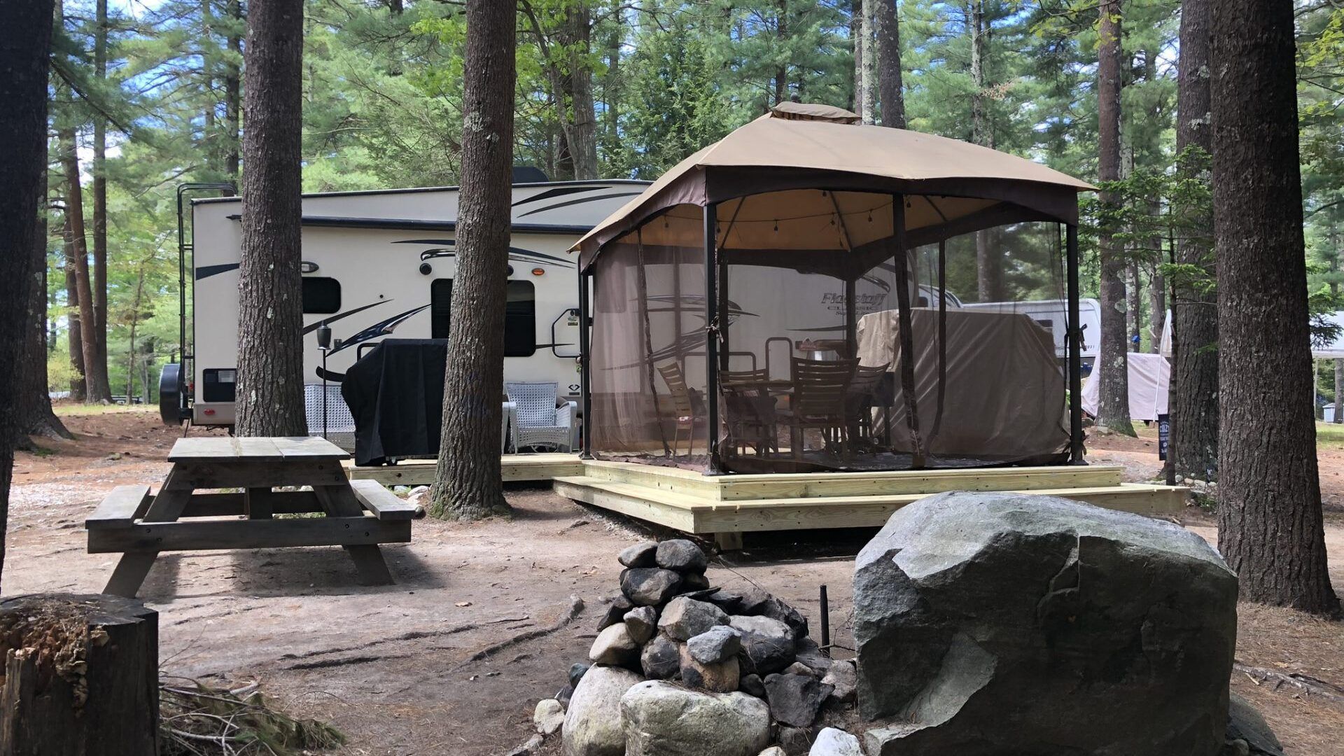 A gazebo in the middle of a forest next to a picnic table and a fire pit.