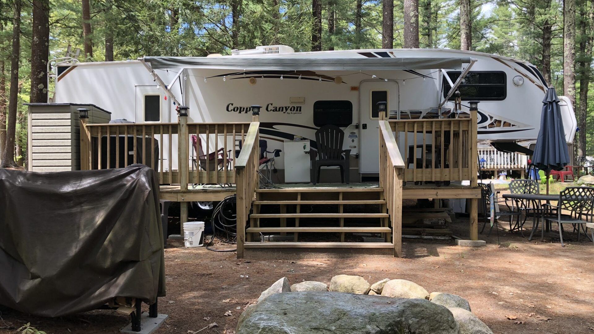 A rv is parked in the woods with a deck and stairs.