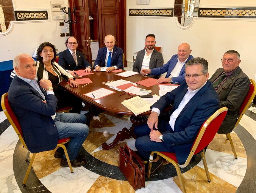 Gruppo di persone sedute attorno a un tavolo, intente a firmare documenti. Stanza riccamente decorata con pavimento in marmo.