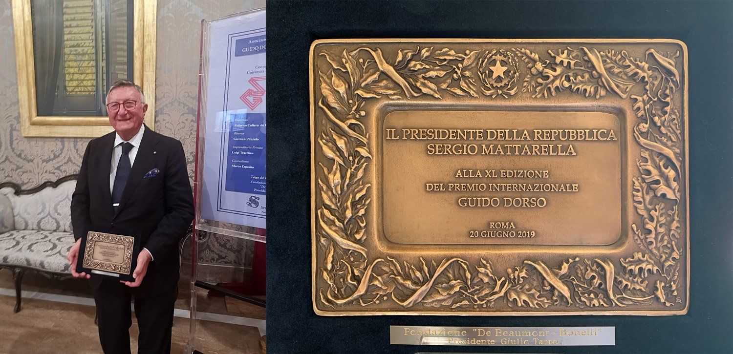 Uomo in giacca e cravatta che regge una targa di premiazione in una sala decorata. Targa di bronzo esposta.