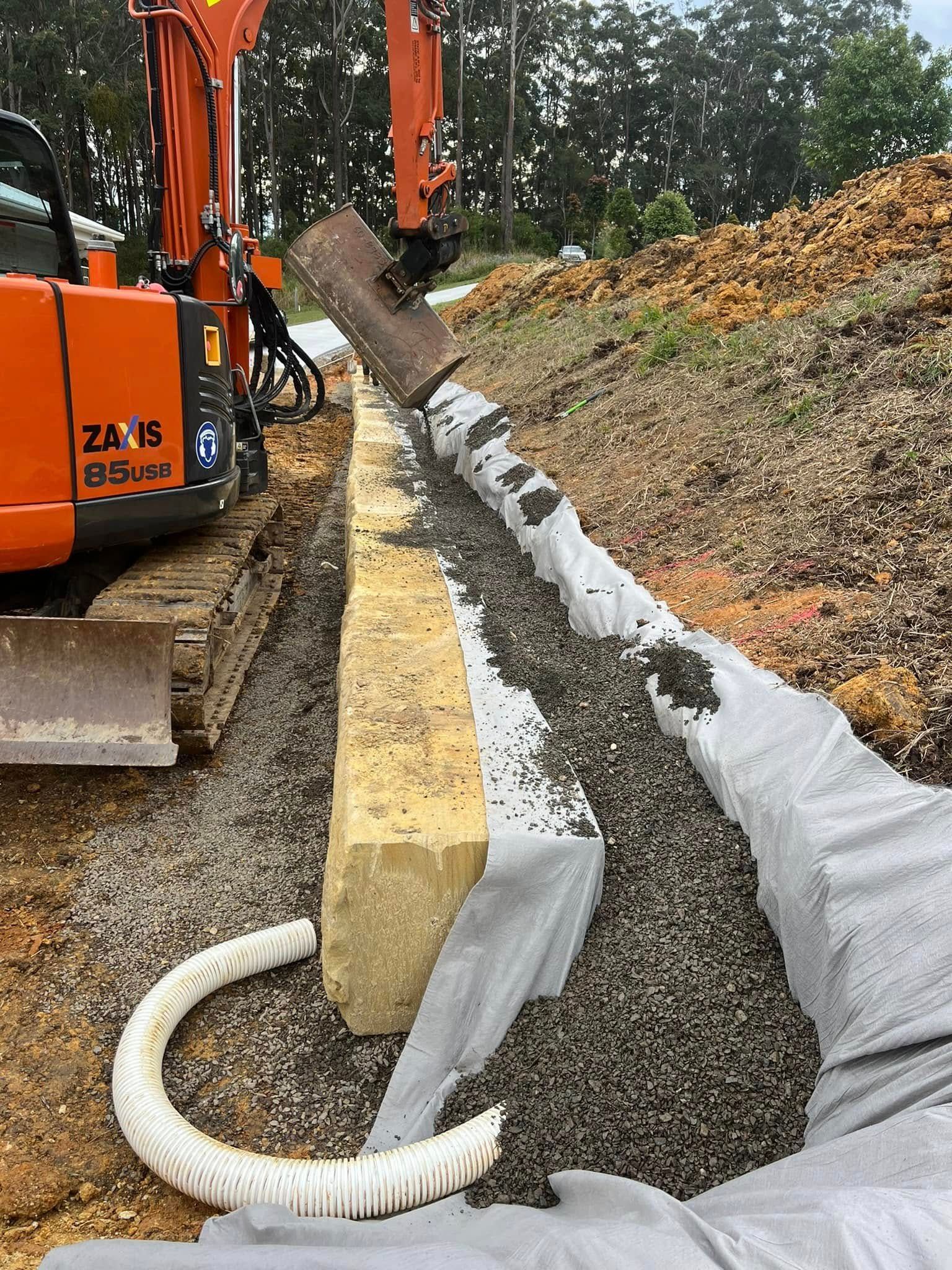 Orange Excavator Laying Drainage Pipe in a Trench Along a Hillside — MG Earthworks and Plant Hire in Nambucca, NSW