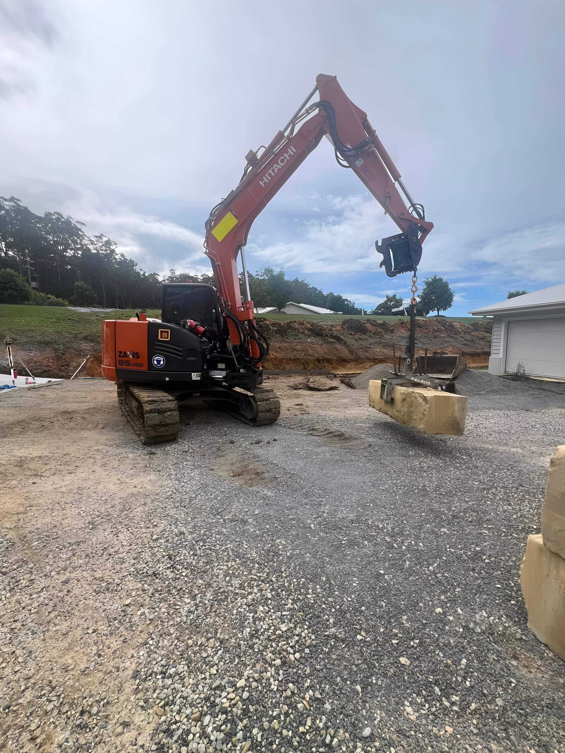 An Excavator Lifts a Large Stone Block on a Construction Site With Gravel — MG Earthworks and Plant Hire in Macksville, NSW