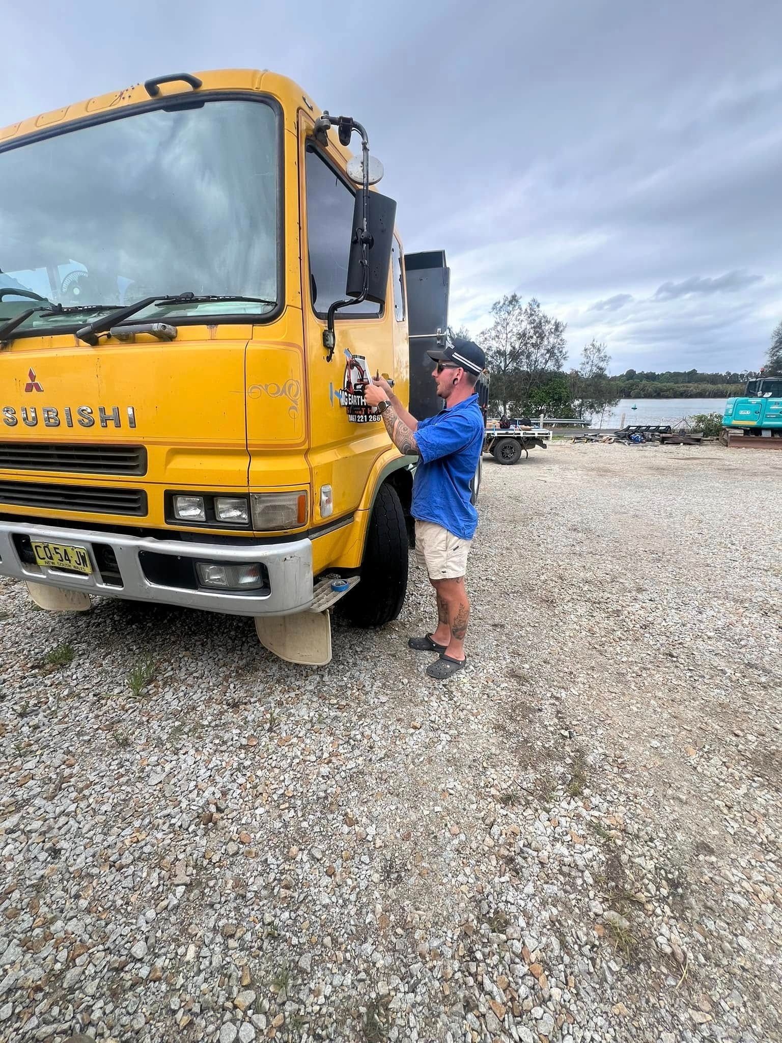 Man Adjusting a Yellow Mitsubishi Truck Mirror on Gravel — MG Earthworks and Plant Hire in Port Macquarie, NSW