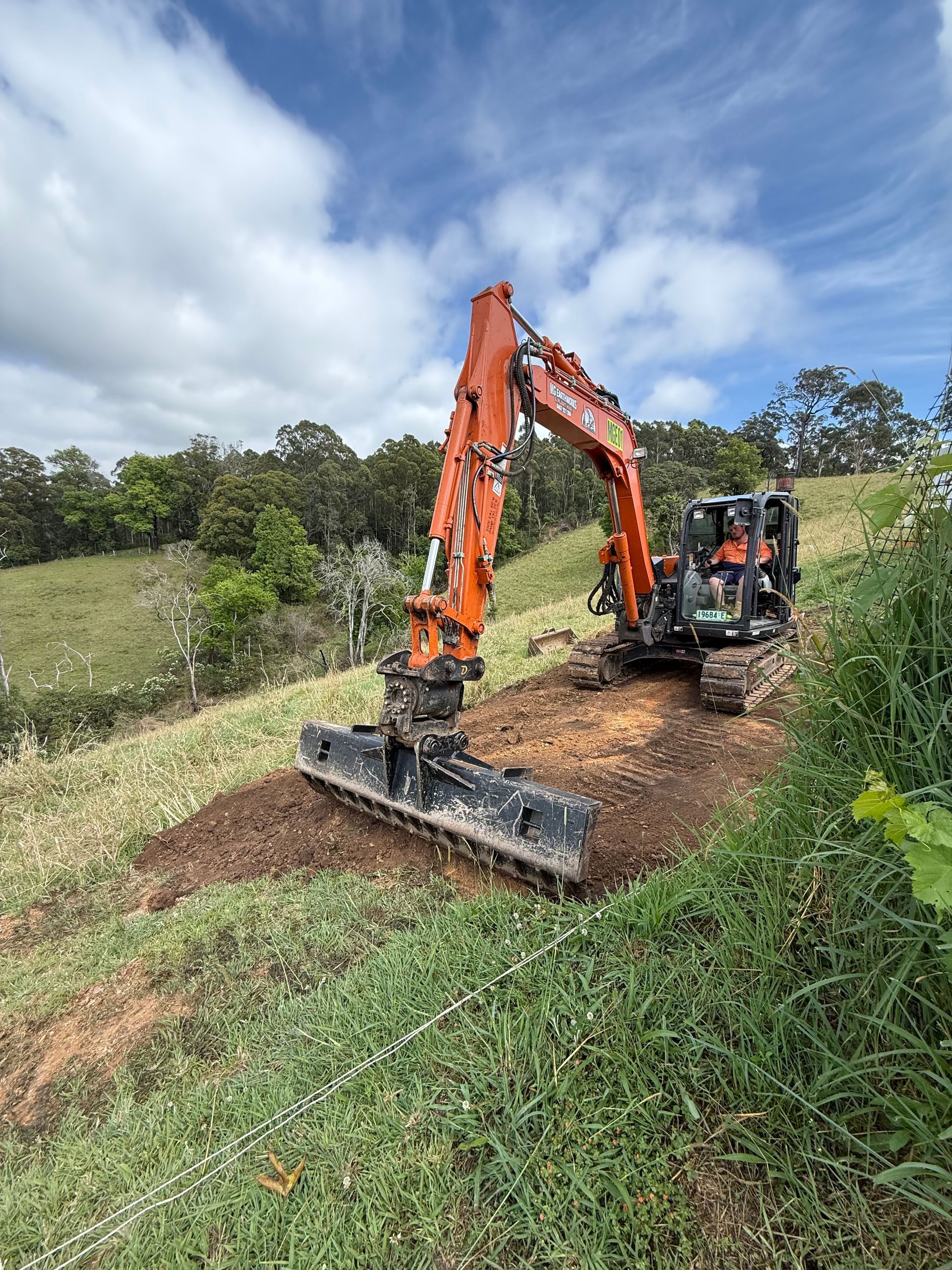 Orange excavator on a grassy hillside levelling dirt with its attachment; blue sky and trees in the background.