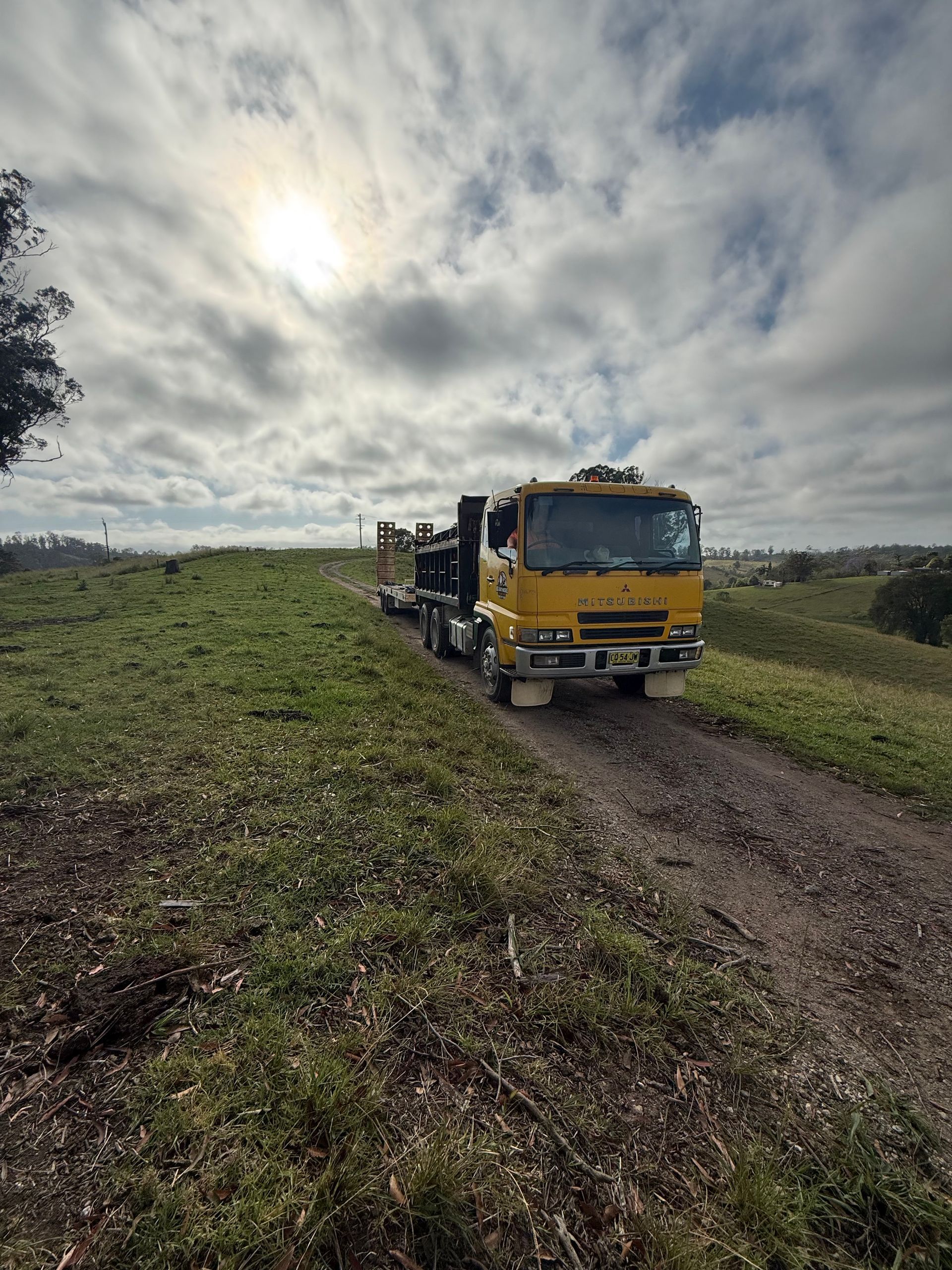 Yellow truck driving on a dirt road, grassy field, cloudy sky.