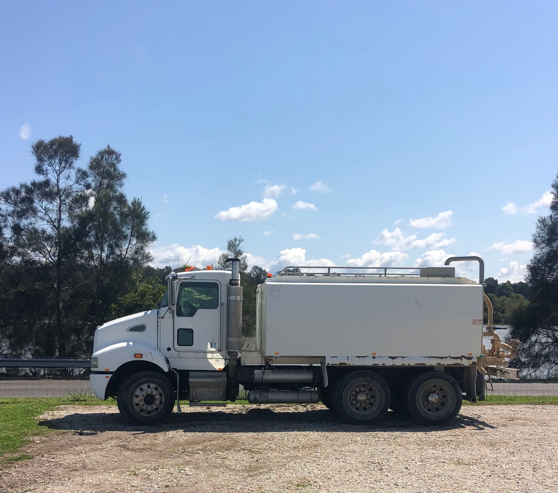 White water tanker truck parked on gravel under a blue sky with trees in the background.