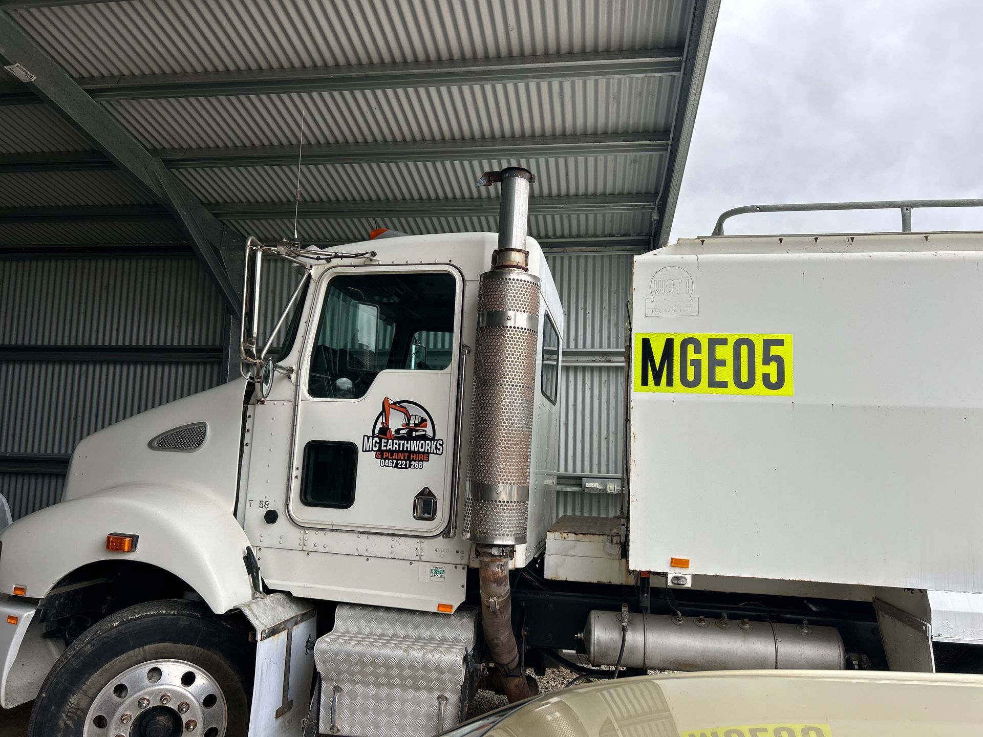 White semi-truck parked under a corrugated metal roof. A yellow license plate reads 