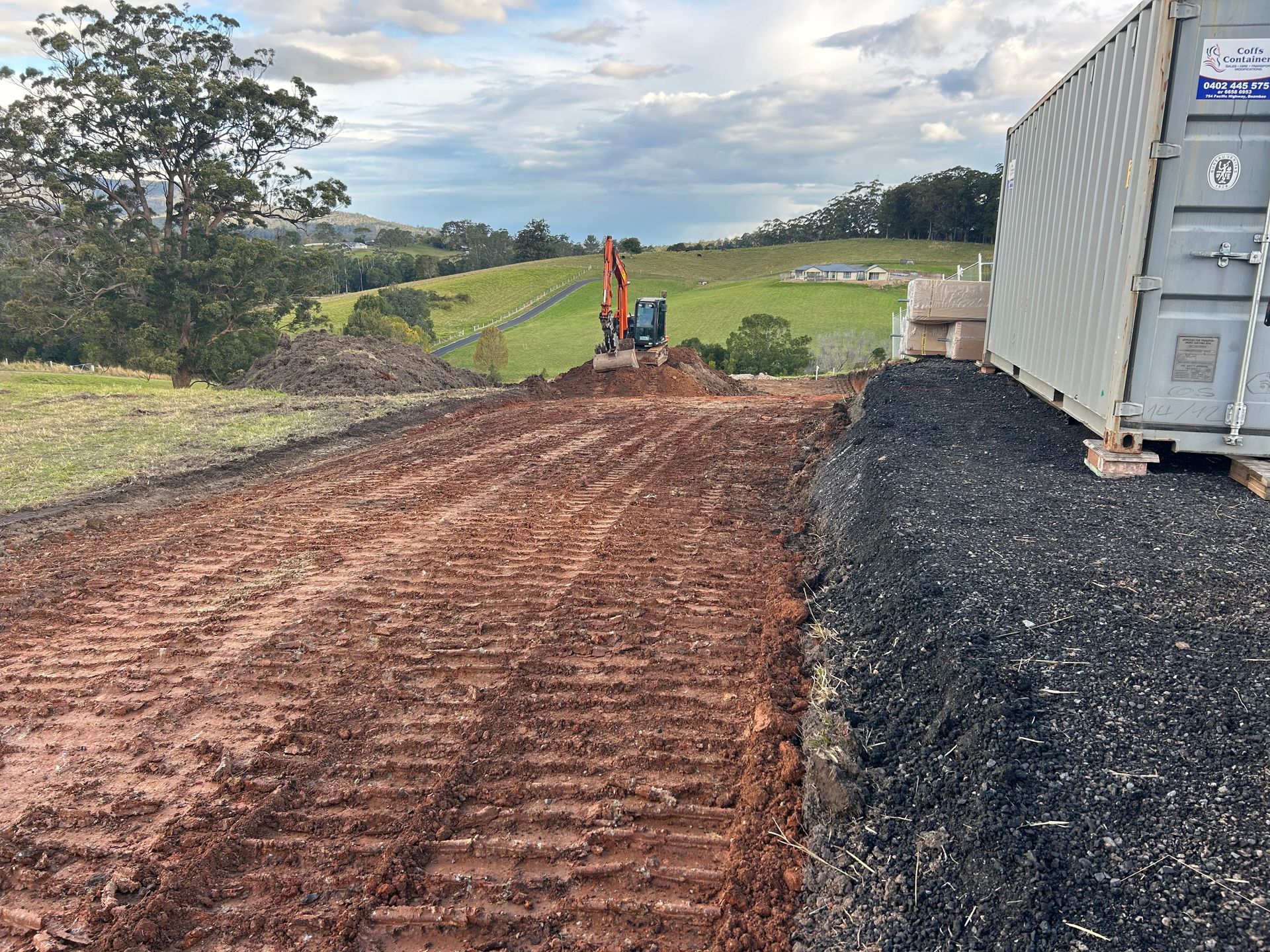Dirt road under construction with excavator, container, and green hills in the distance.