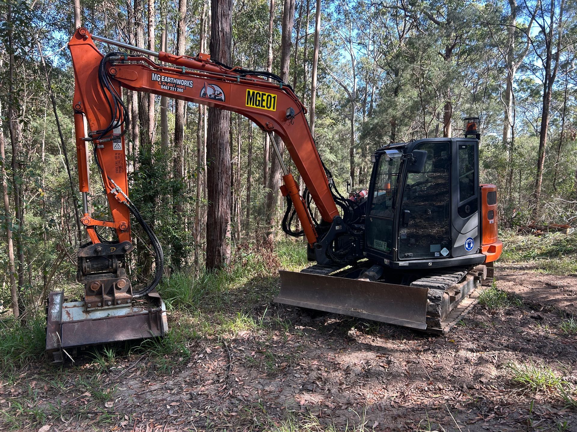 Orange excavator with a brush cutter attachment in a wooded area.