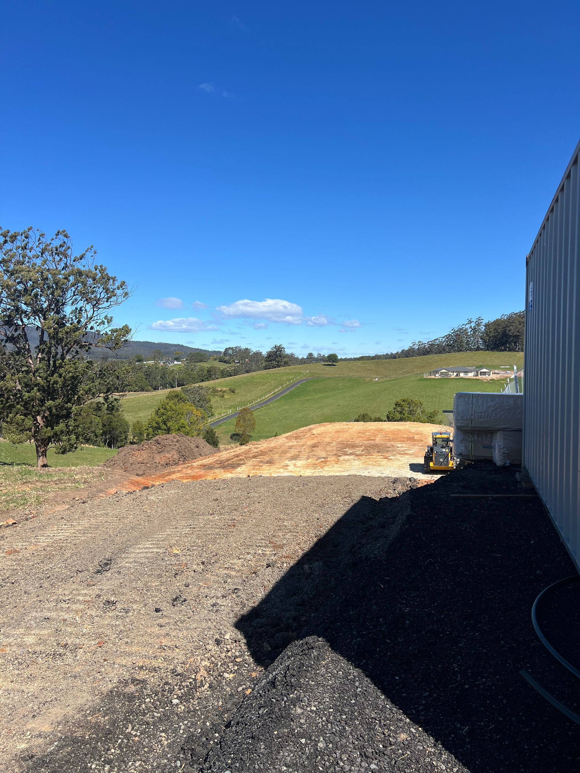 A sunny view of a grassy hill and blue sky. Construction site with gravel and a building in the frame.
