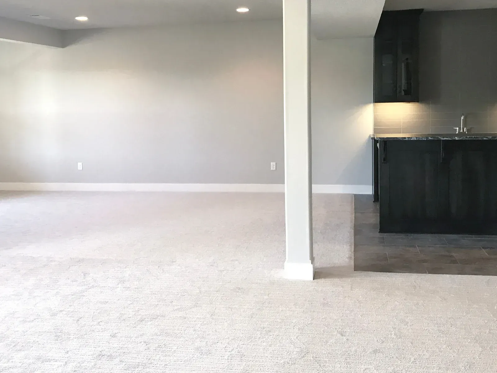 An empty room with a carpeted floor and a kitchen in the background.