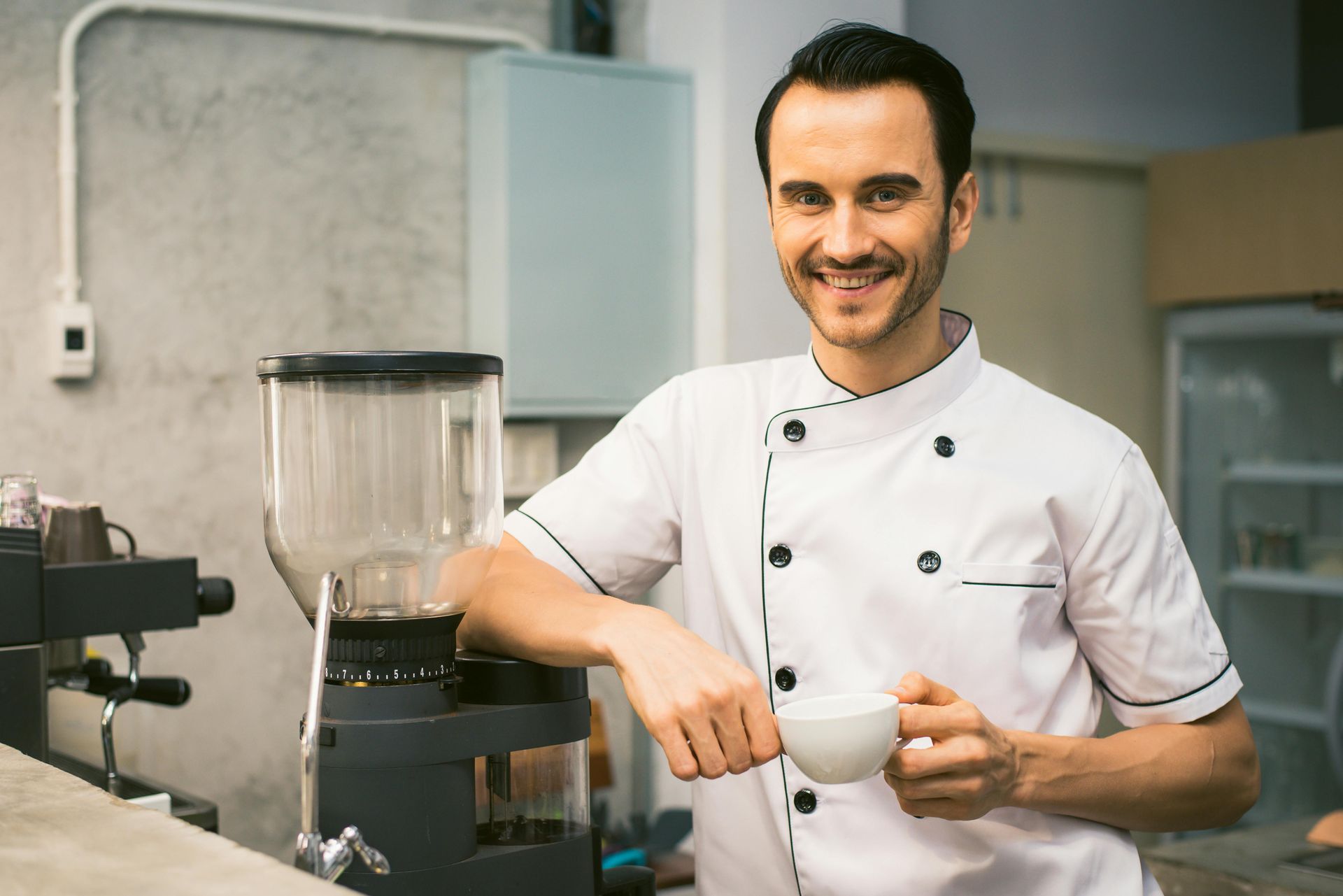 Barista smiling in a white uniform pouring coffee beside a grinder in a cafe kitchen