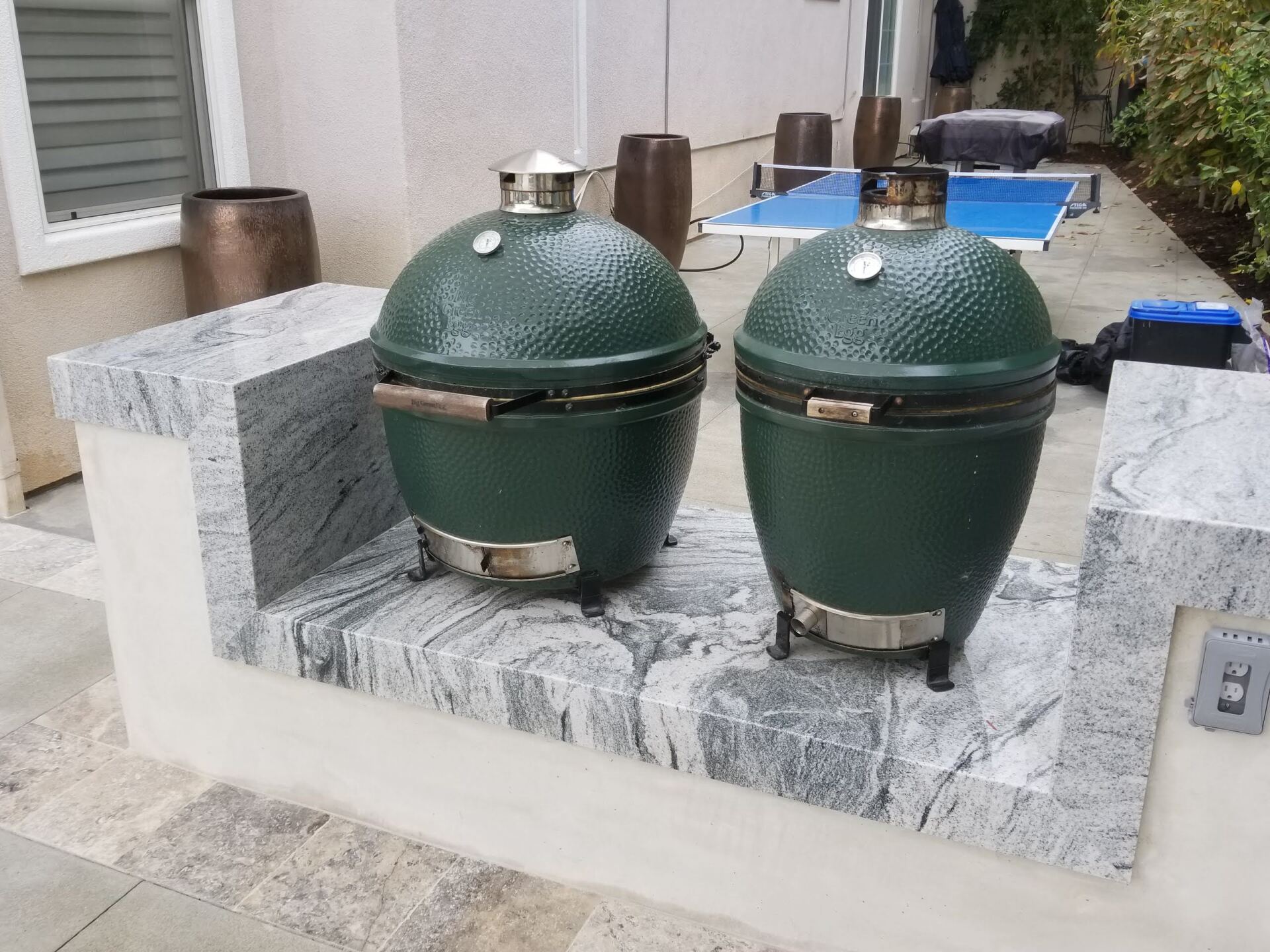 Two green ceramic grills on a granite ledge, outdoors with a table and containers in the background.