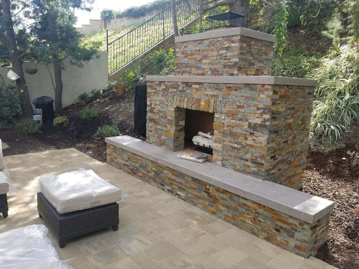 Outdoor stone fireplace with seating area; surrounded by landscaping and a hillside.