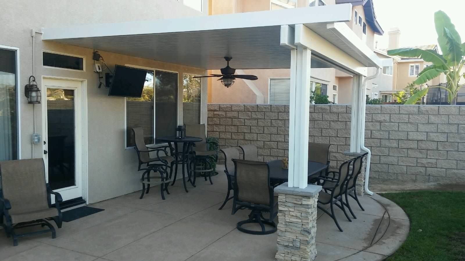 Patio with a covered seating area, featuring tables, chairs, a TV, and a ceiling fan; adjacent to a brick wall.