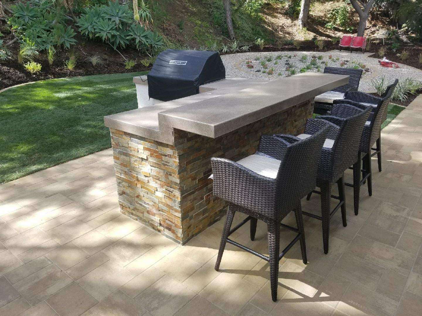 Outdoor bar with grill, stone facade, concrete countertop, and wicker bar stools on a patio.