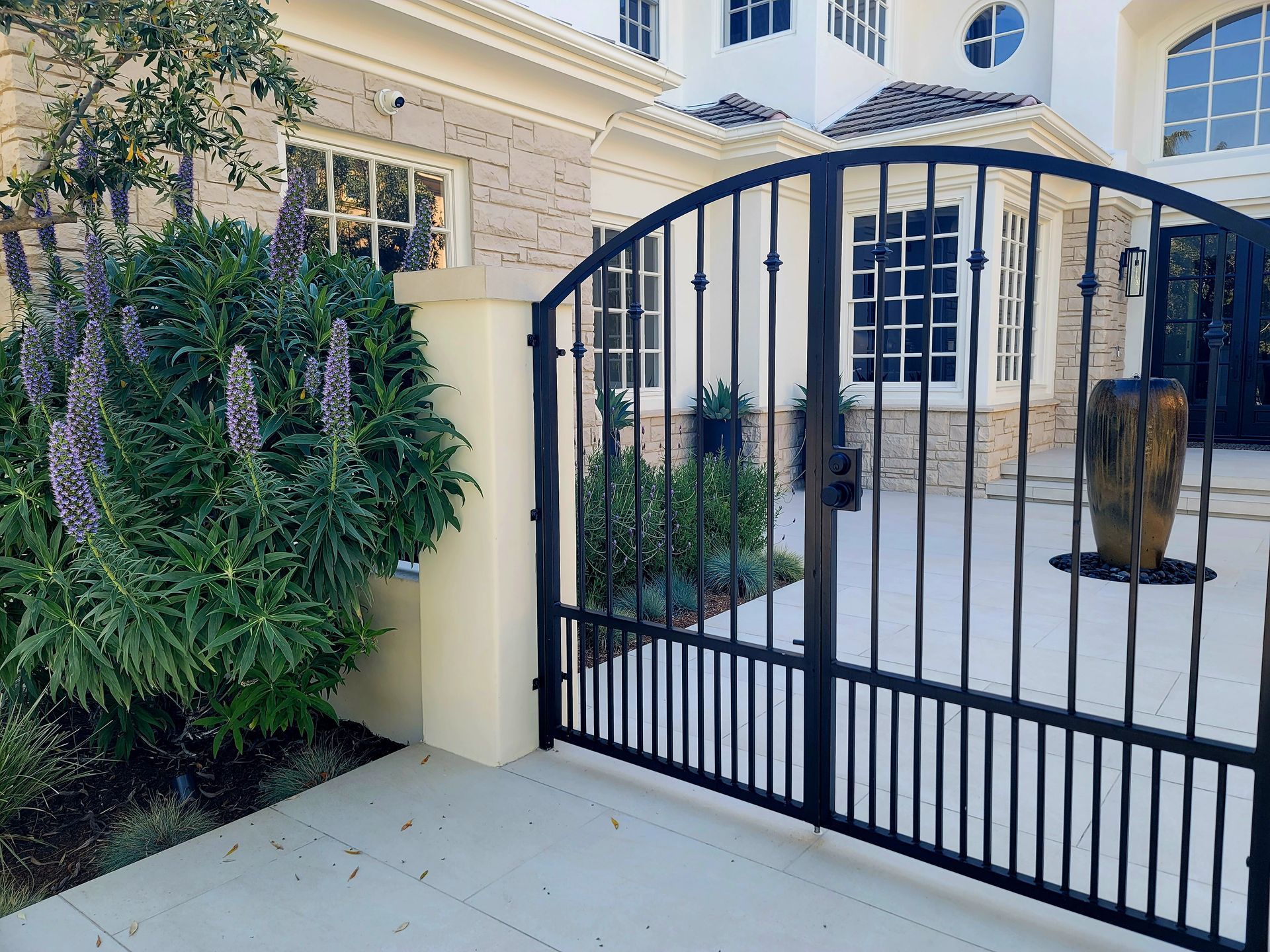 Black wrought-iron gate in front of a stone-clad house, with purple-flowered bush to the left.