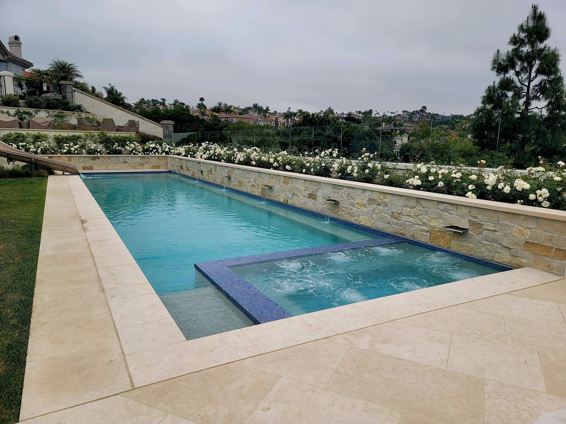 Rectangular pool with a spa, surrounded by stone and flowers, under an overcast sky.