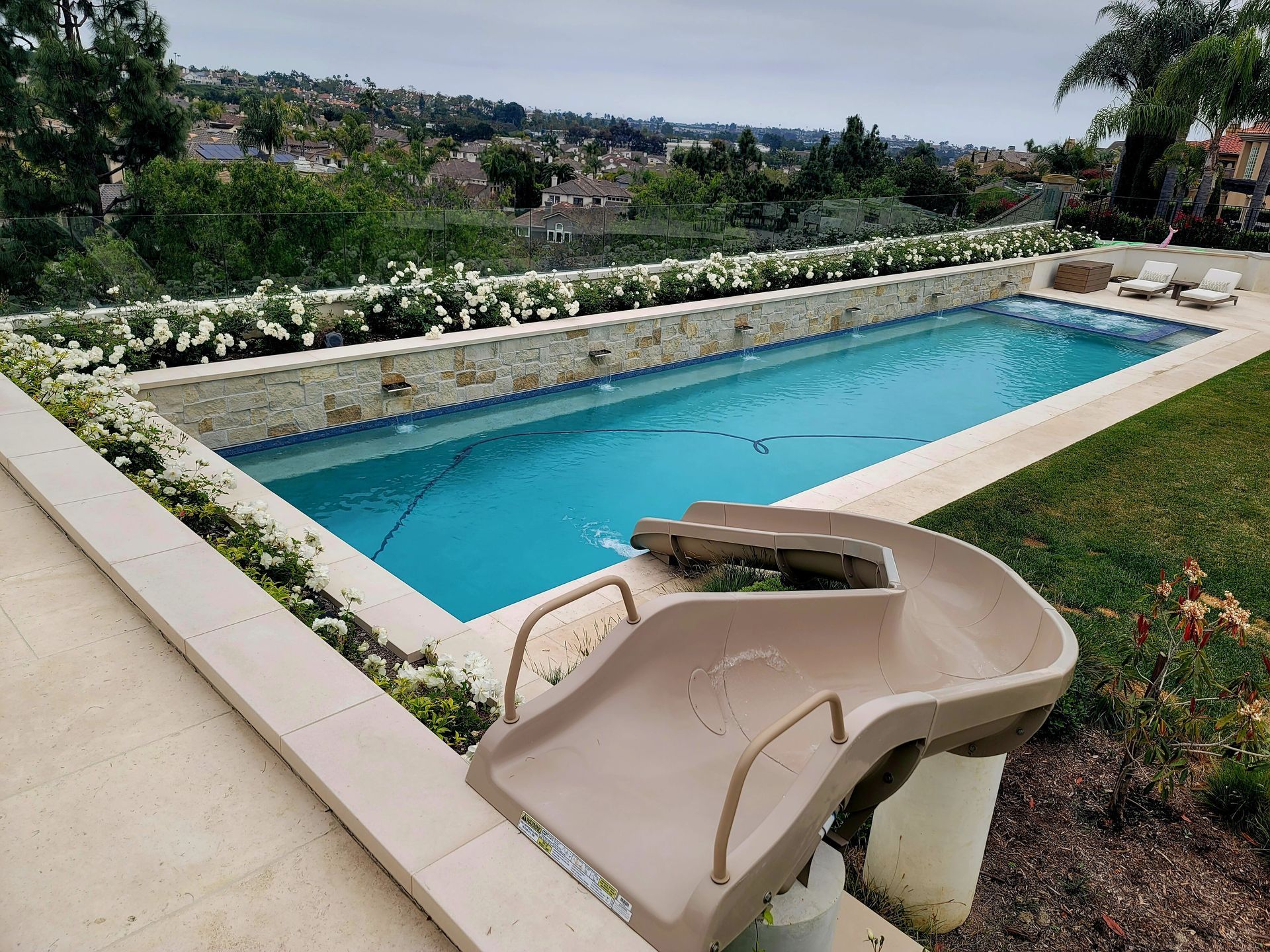 A long, rectangular pool with a slide, surrounded by greenery and a hillside view.