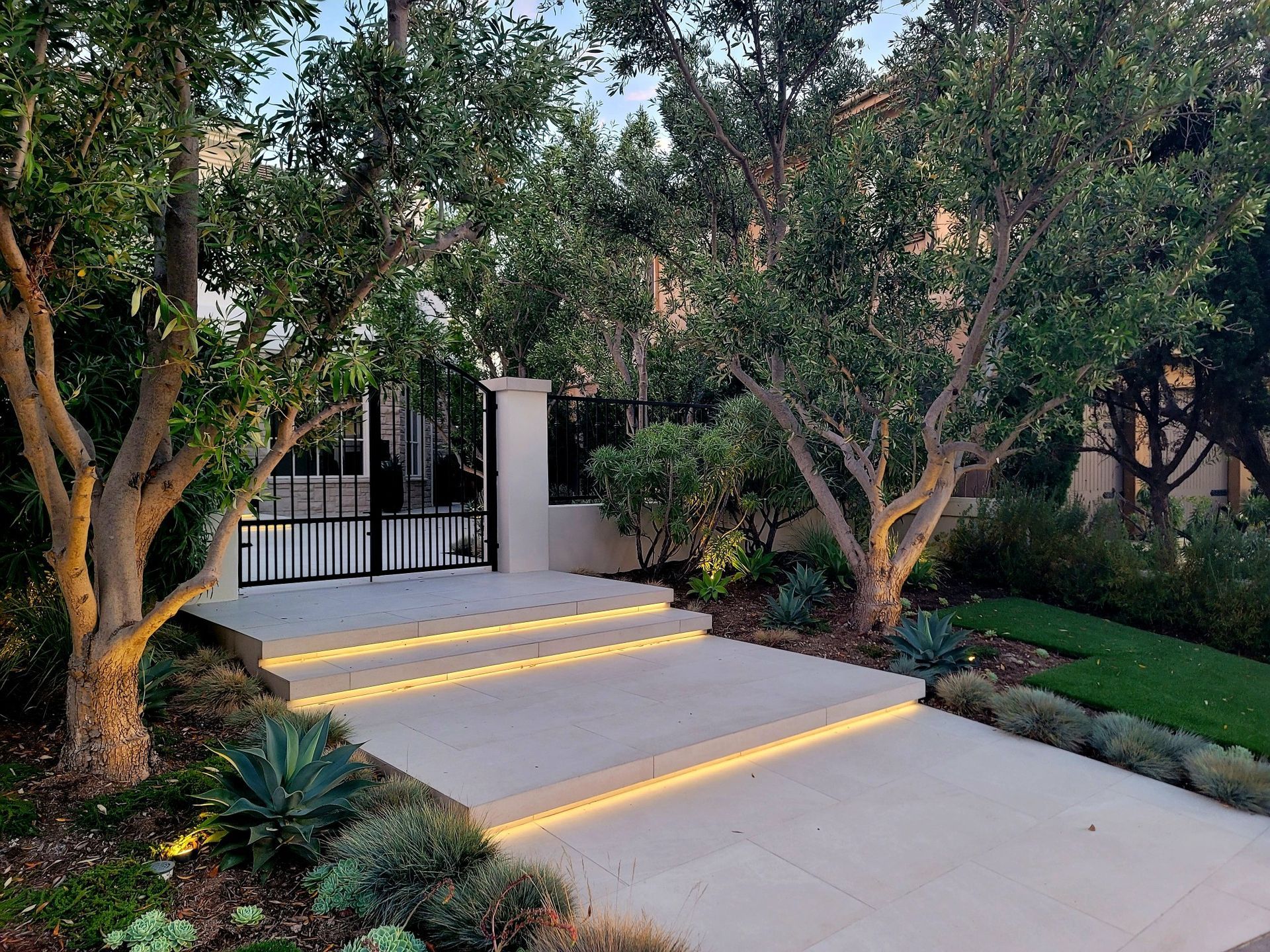 Stone steps with strip lighting leading to a gate, flanked by trees and greenery.