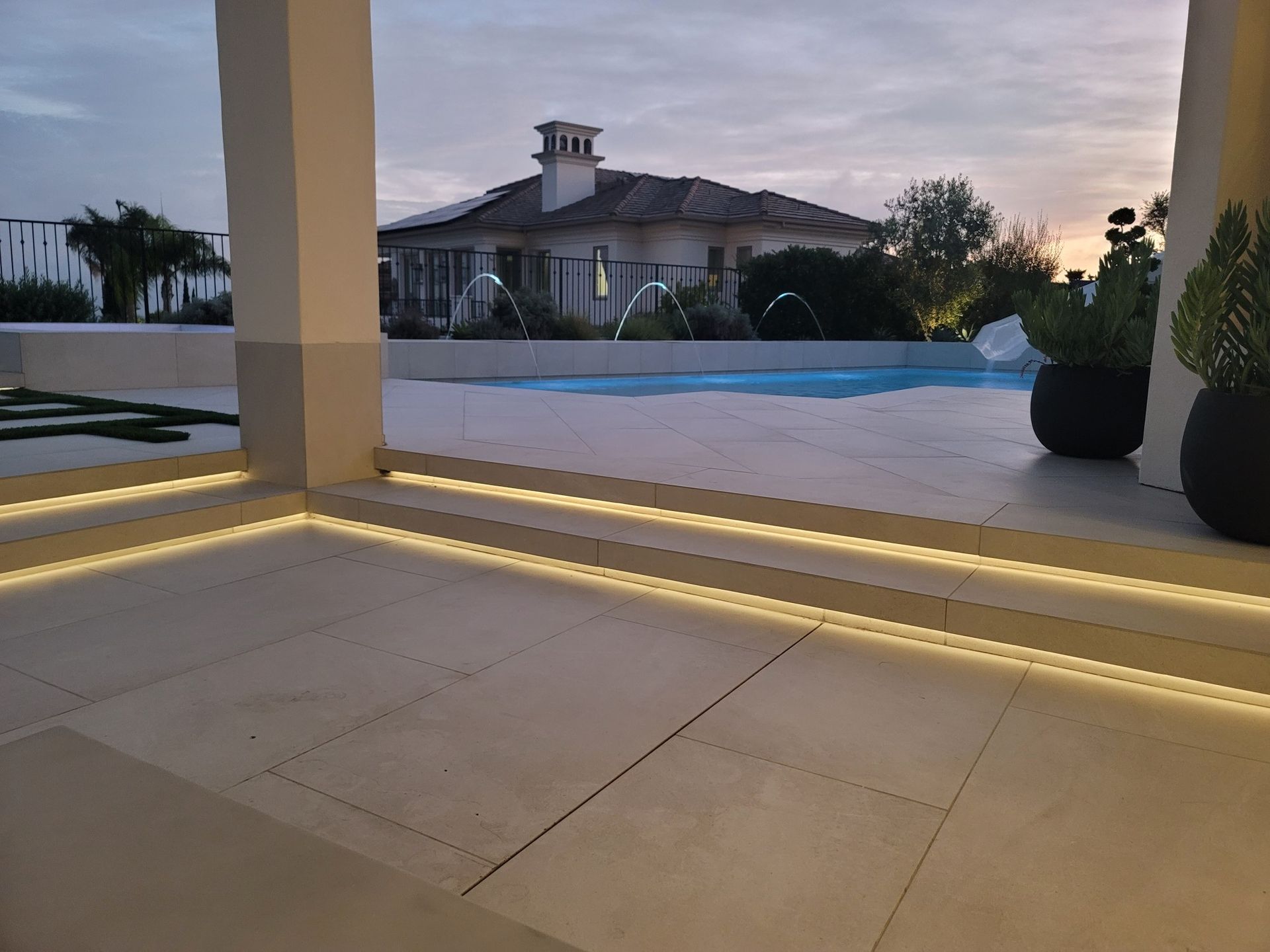 Patio with illuminated edges overlooking a pool and a house at dusk.