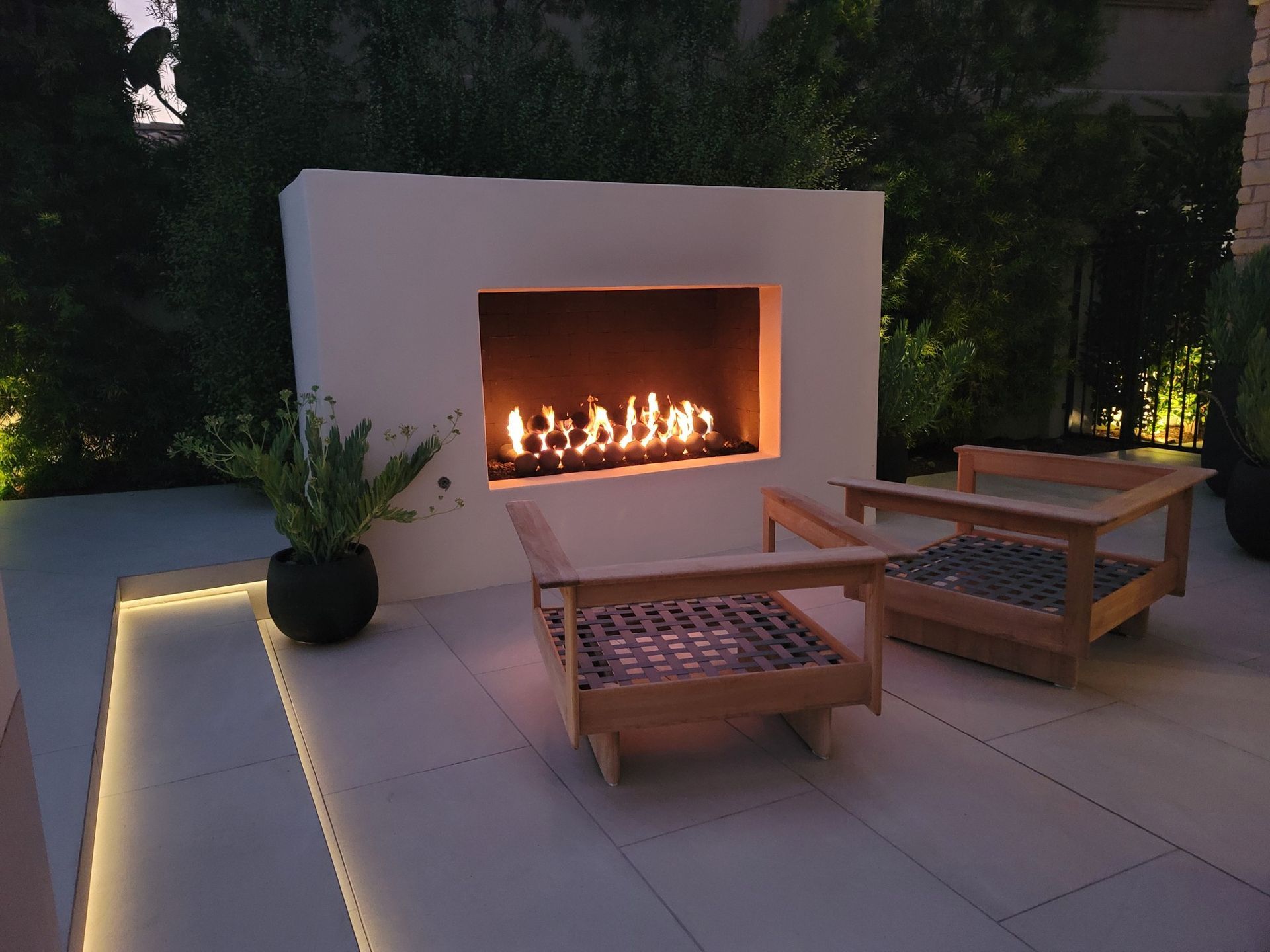 Outdoor fireplace with two wooden chairs, lit fire. Evening setting with ambient lighting.