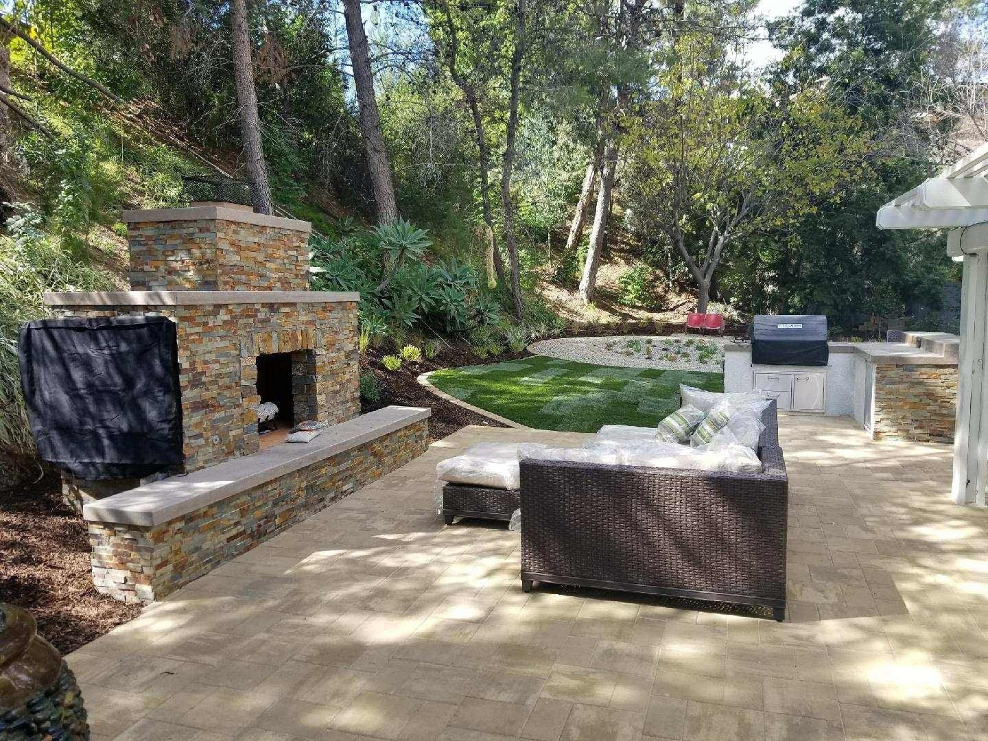 Outdoor patio with stone fireplace, seating, and grill, surrounded by trees and greenery.