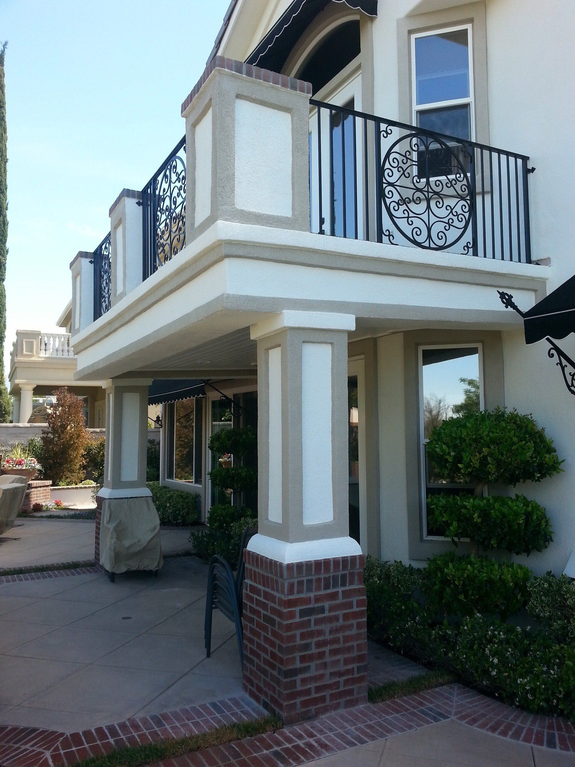 Exterior of a building with a balcony. Beige and white walls, brick pillars. Black wrought iron railing. Sunny day.