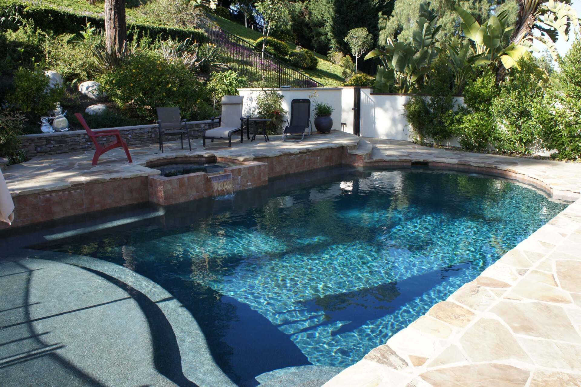 Swimming pool with blue water and stone deck in a lush green yard. Chairs and trees visible.