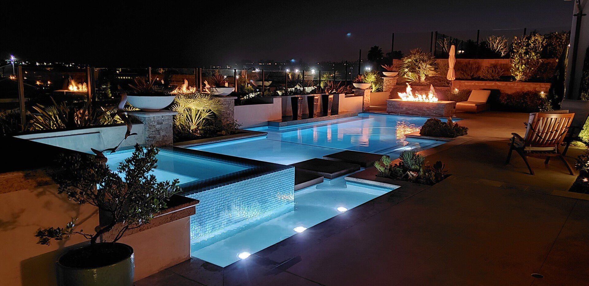 Nighttime view of a lit pool and jacuzzi area with fire pit, trees, and city lights in the background.