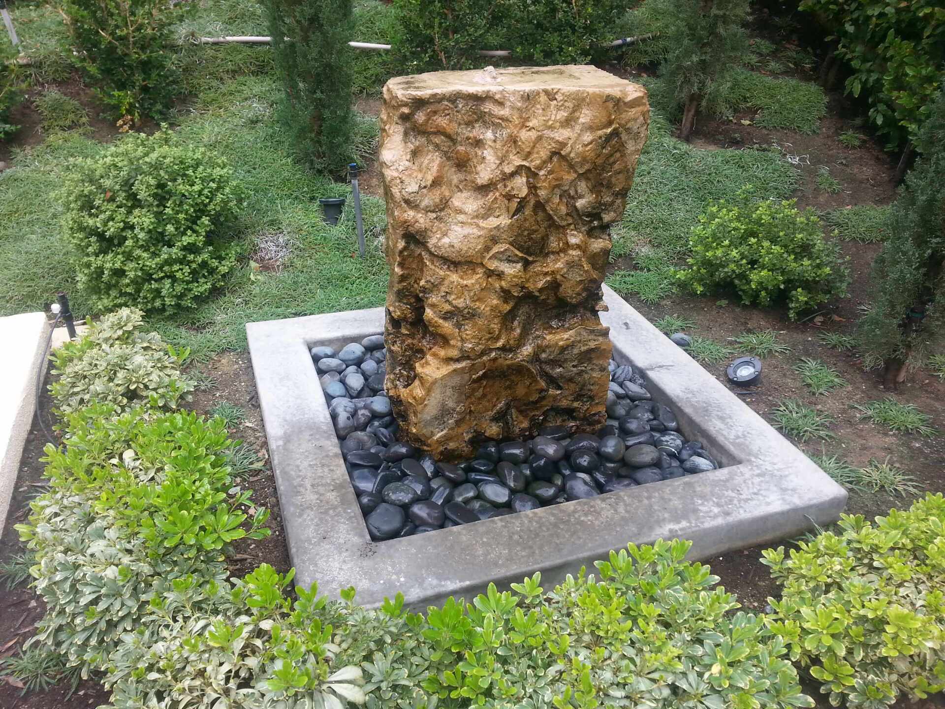 Stone fountain with black pebbles, surrounded by greenery in a garden setting.