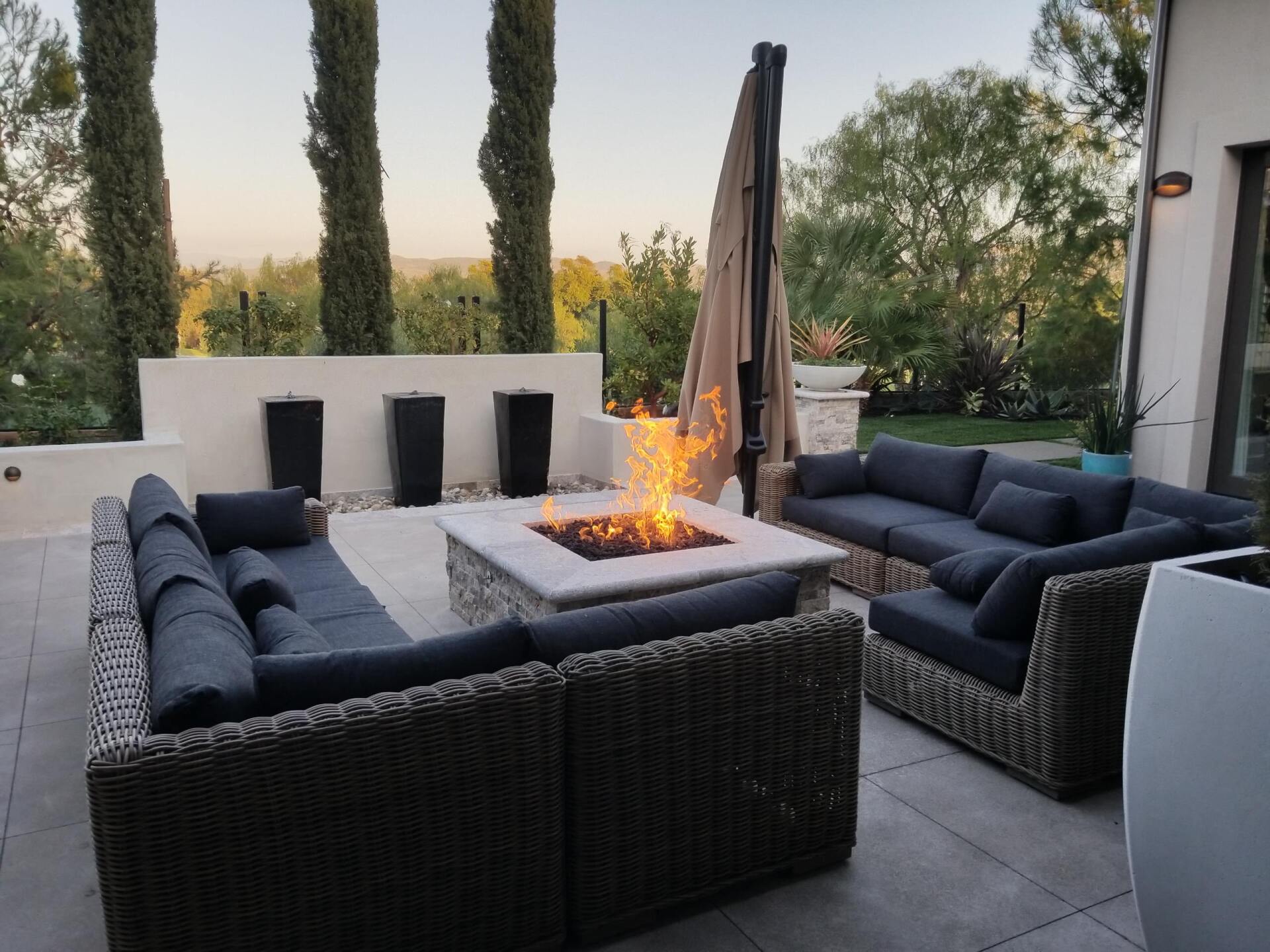 Outdoor patio with fire pit, sectional sofa, and decorative plants. Sunset background.