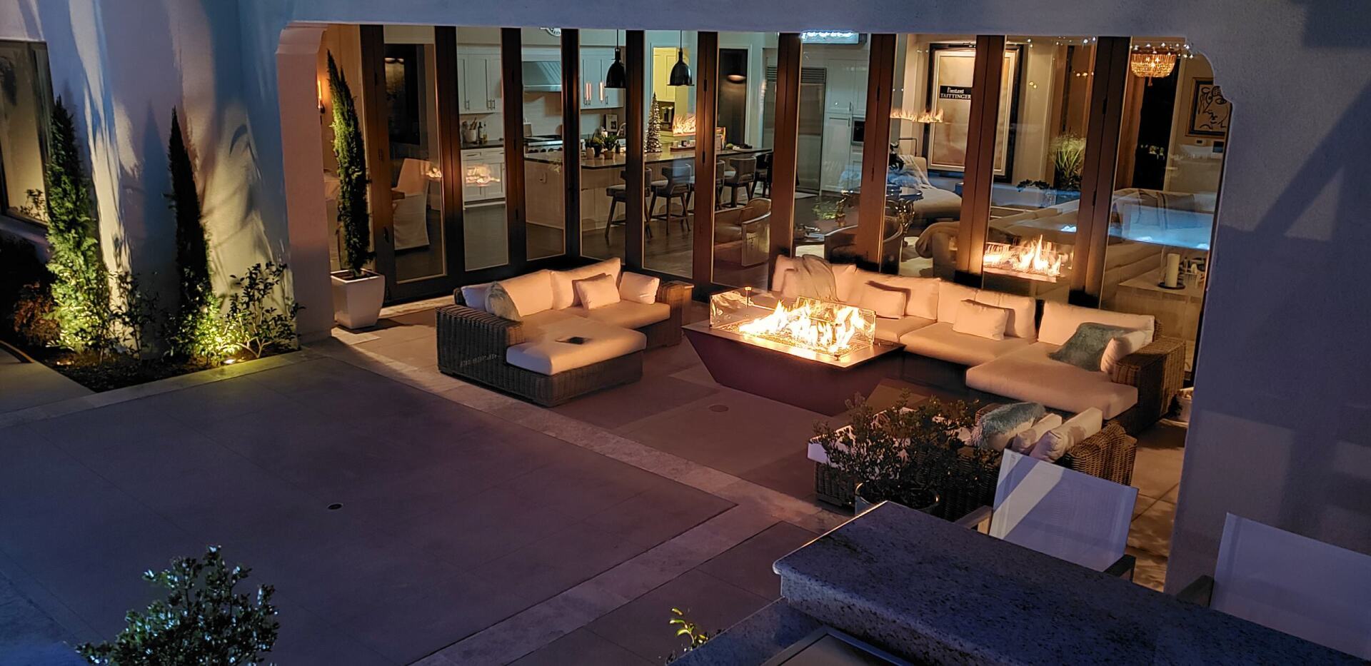 Outdoor living space at night, with lit fire pits and sofas, adjacent to a modern home.