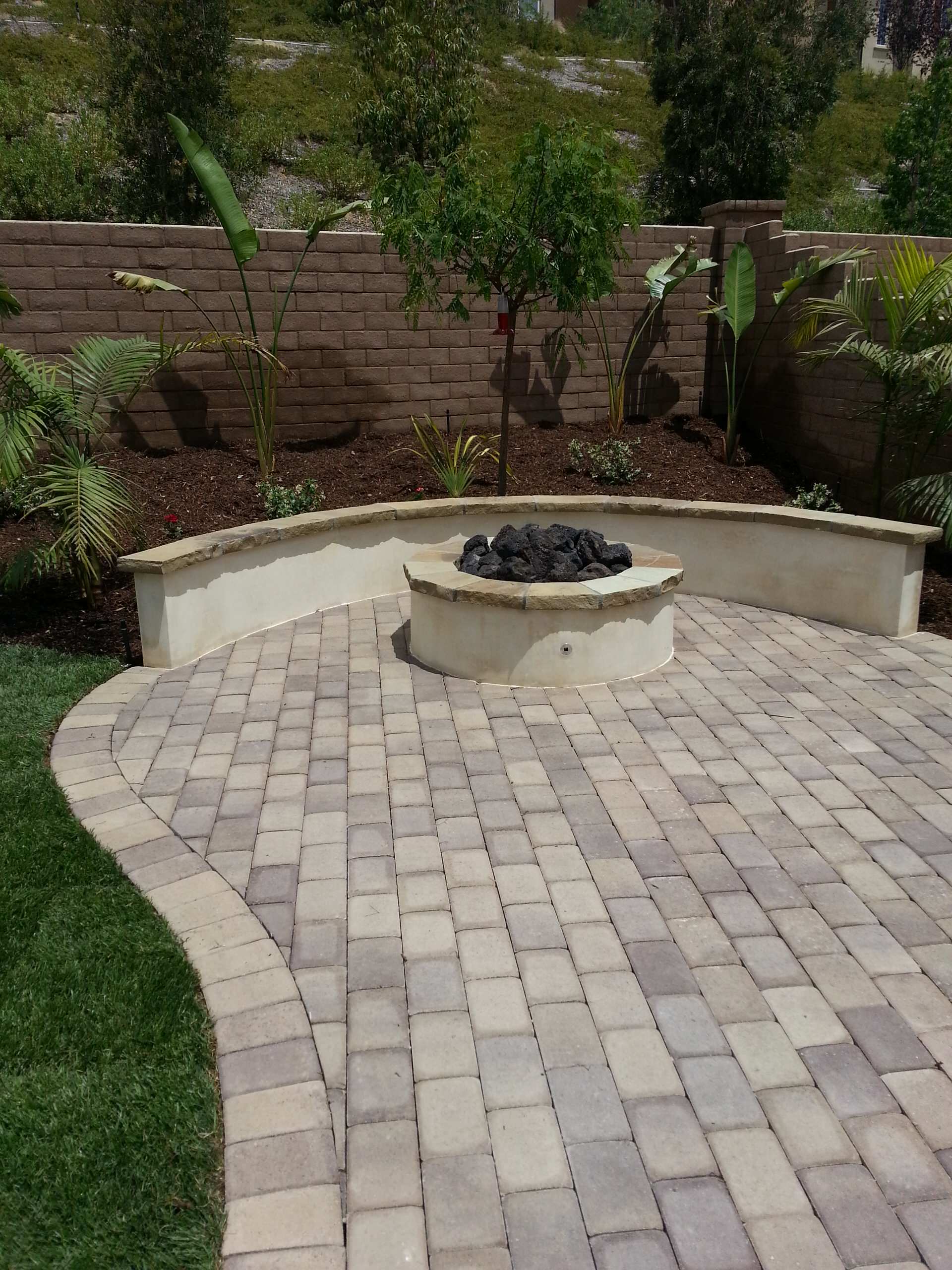 Stone patio with a fire pit, surrounded by a low wall and lush landscaping.