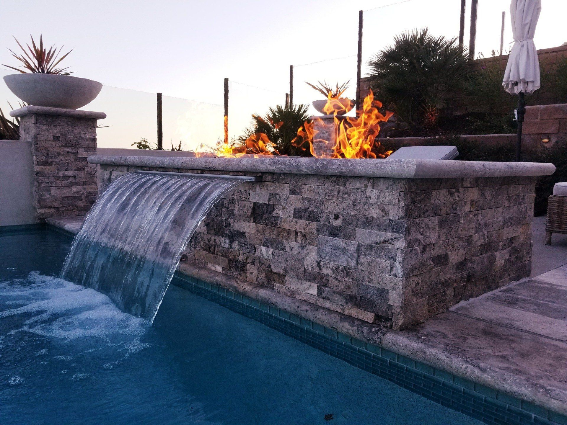 A pool with a stone waterfall and fire feature at dusk.