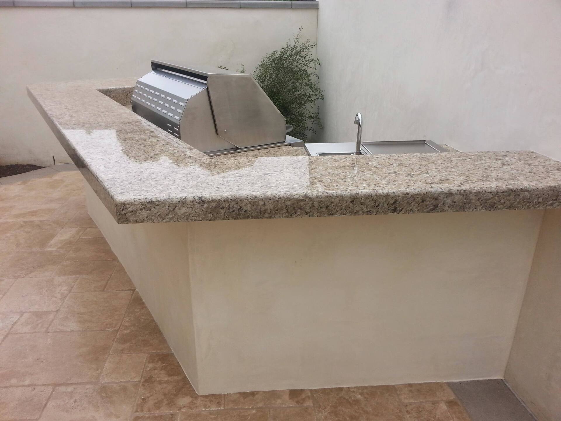Outdoor kitchen with granite countertop, stainless steel grill, and sink on a beige stucco structure.