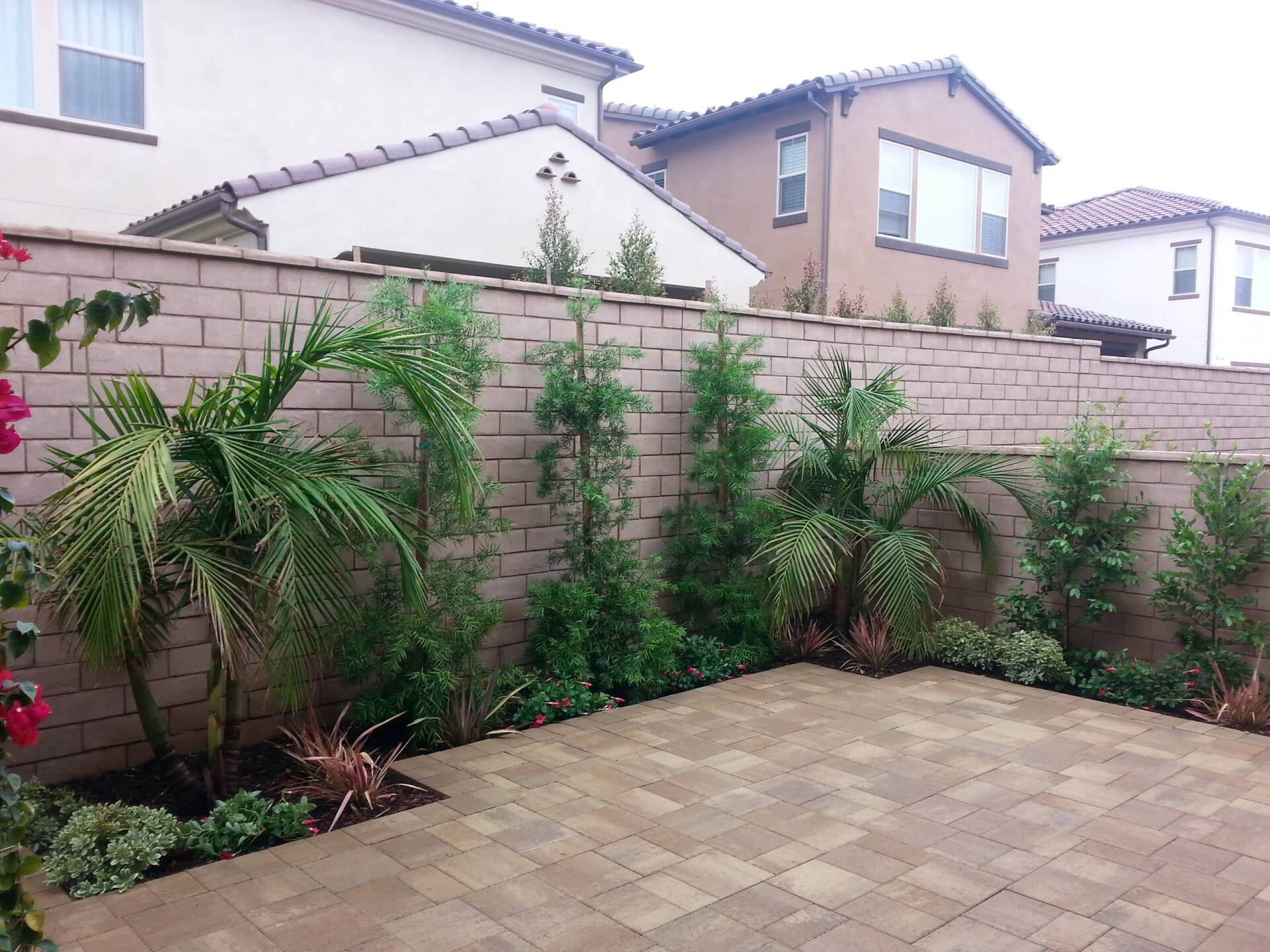 Brick patio with landscaping along a tan brick wall; houses in the background.