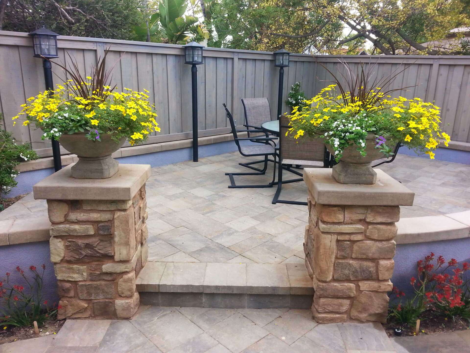 Stone columns with flower pots lead to a paved patio with seating, enclosed by a wooden fence.