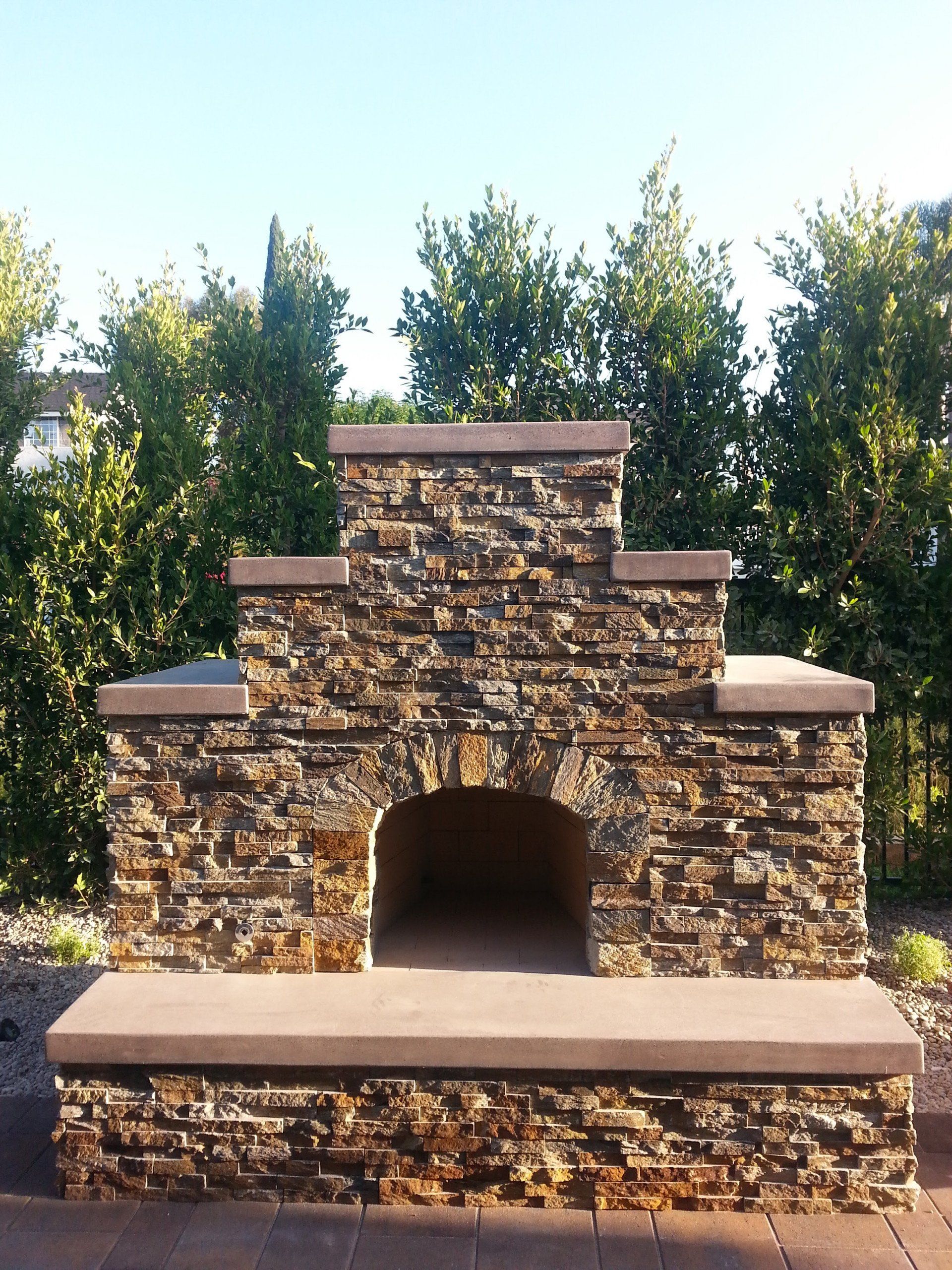 Stone outdoor fireplace with an arched opening, surrounded by greenery under a sunny sky.
