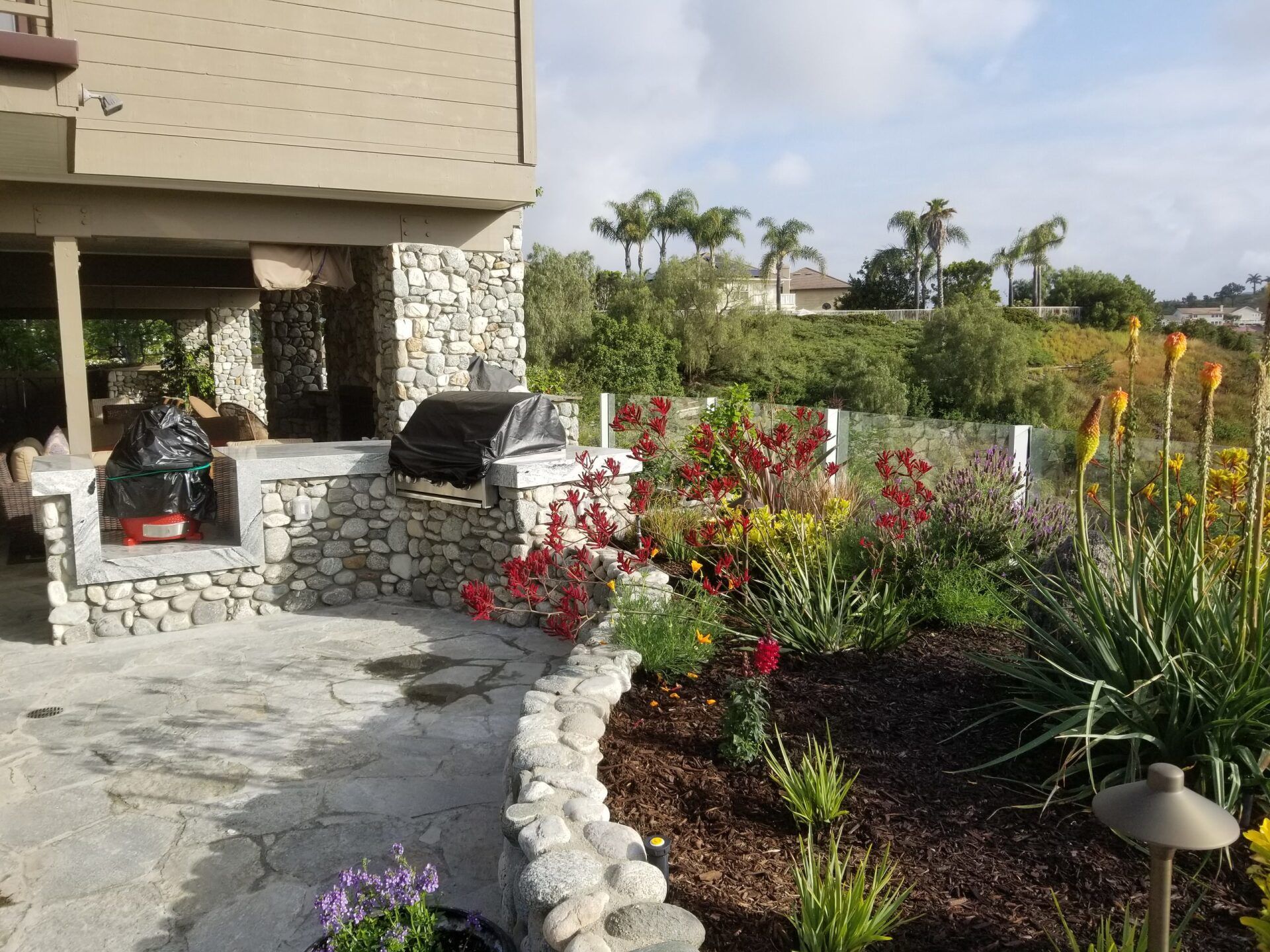Stone patio with built-in grill, lush garden with colorful flowers, house in background.